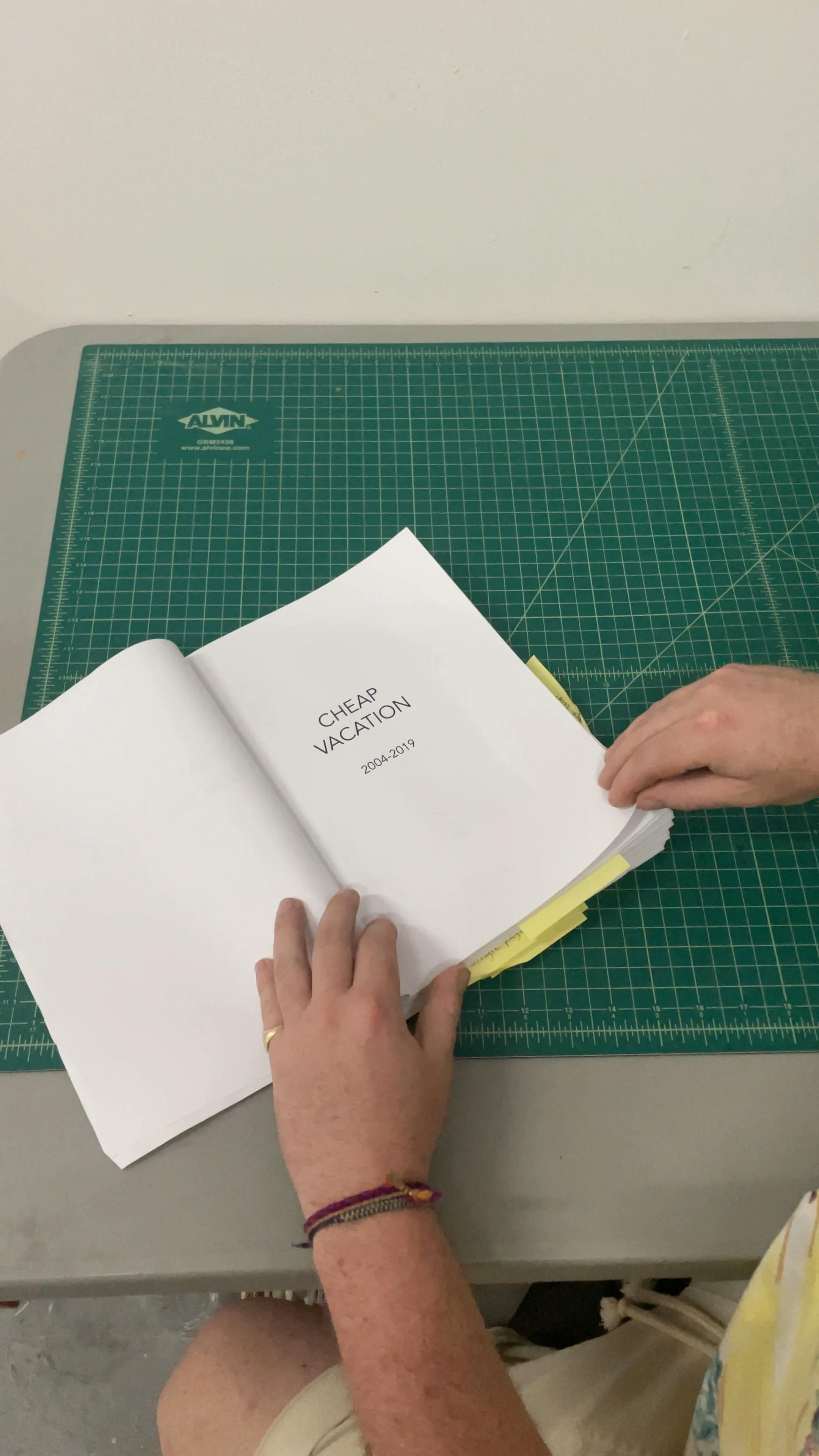 A closeup of artist Ryan James MacFarland flipping through a white manuscript with the title "CHEAP VACATION" on the front.