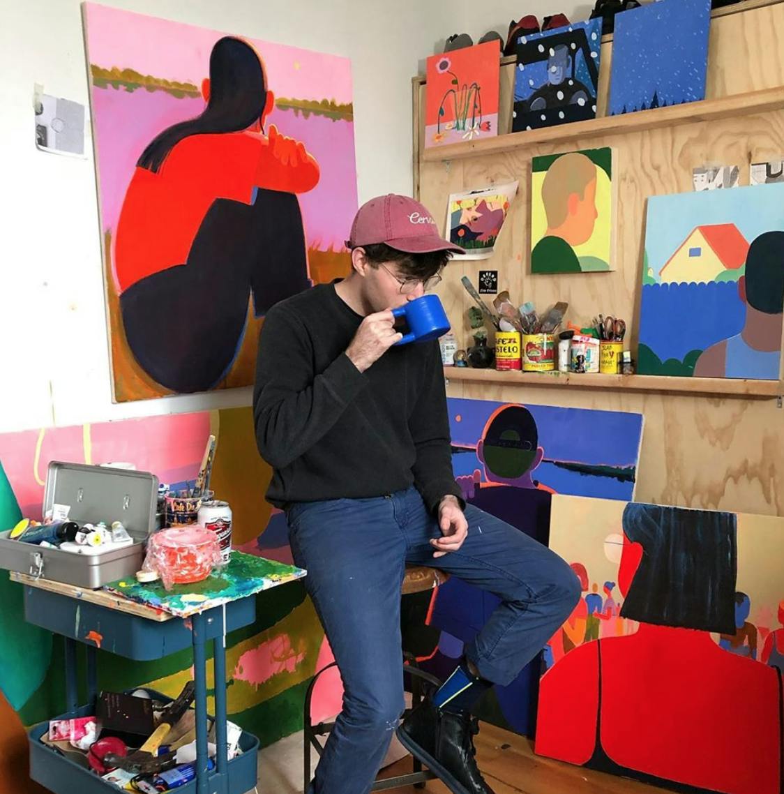 Artist Jackson Joyce surrounded by his paintings in the studio drinking coffee from a blue mug.