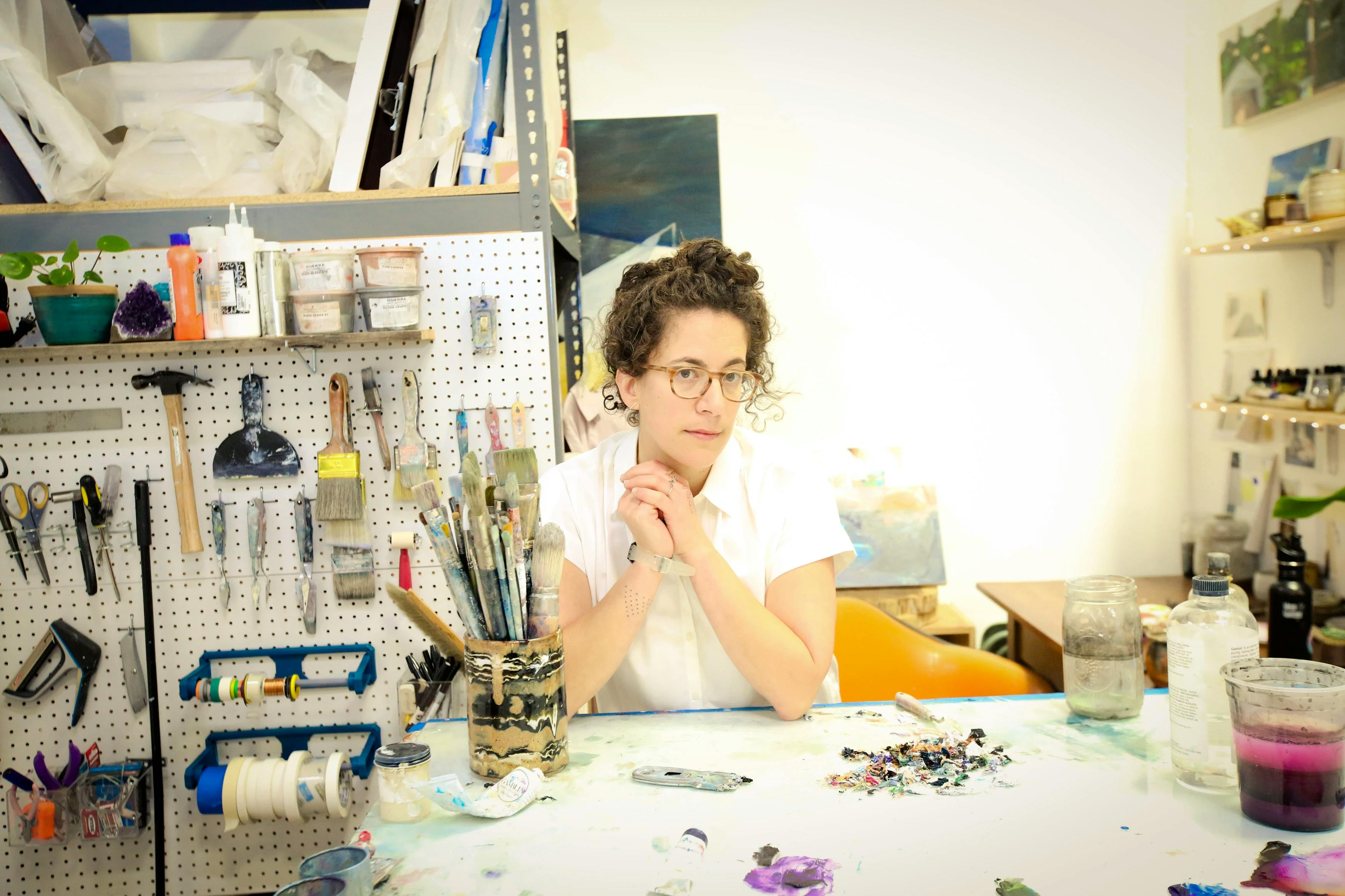 Artist Aliza Cohen wearing a white t-shirt and standing at table inside her studio, surrounded by paintbrushes and other tools.