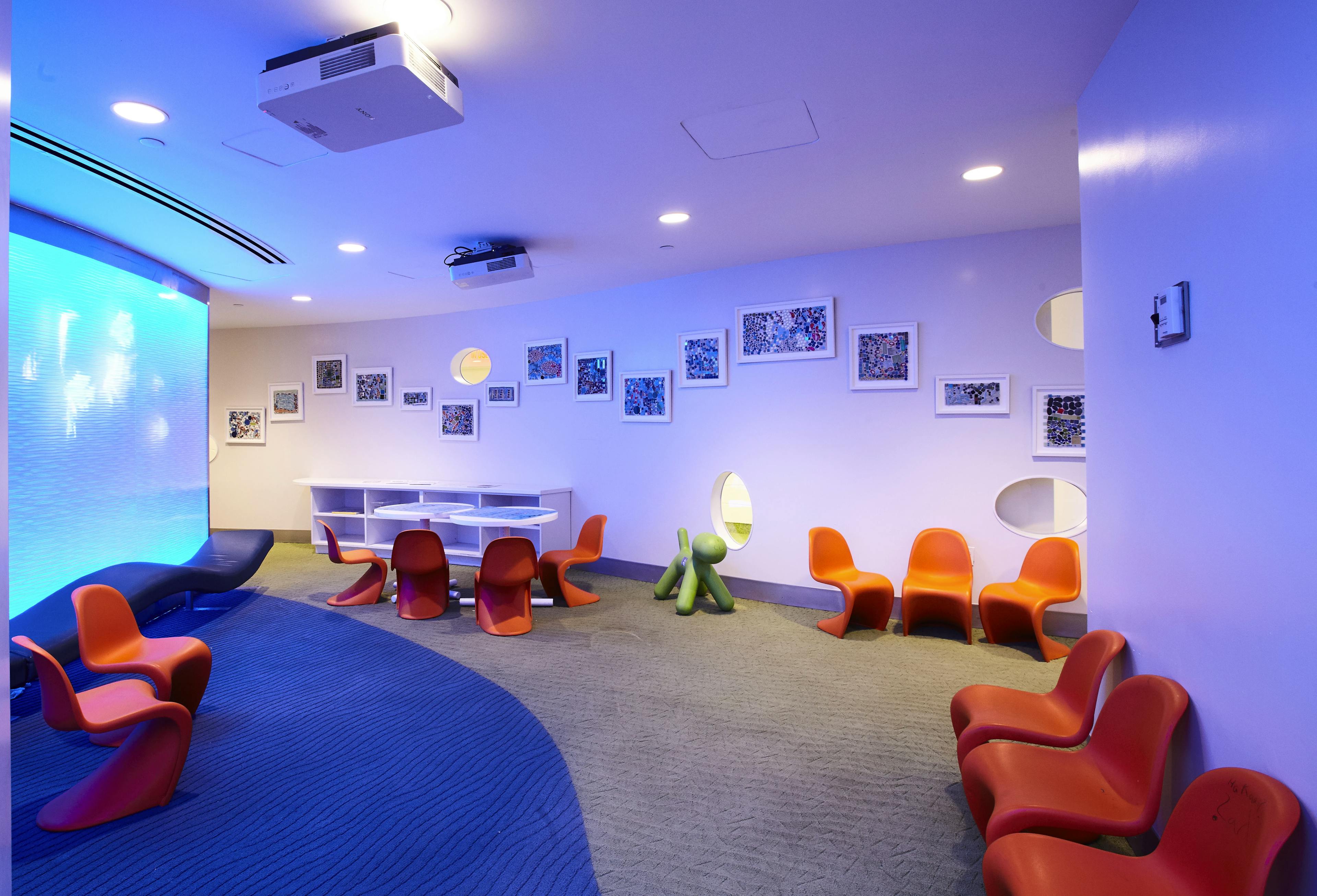 Children's lounge area at a hospital with rows of modern orange chairs and a gallery wall of framed artwork.