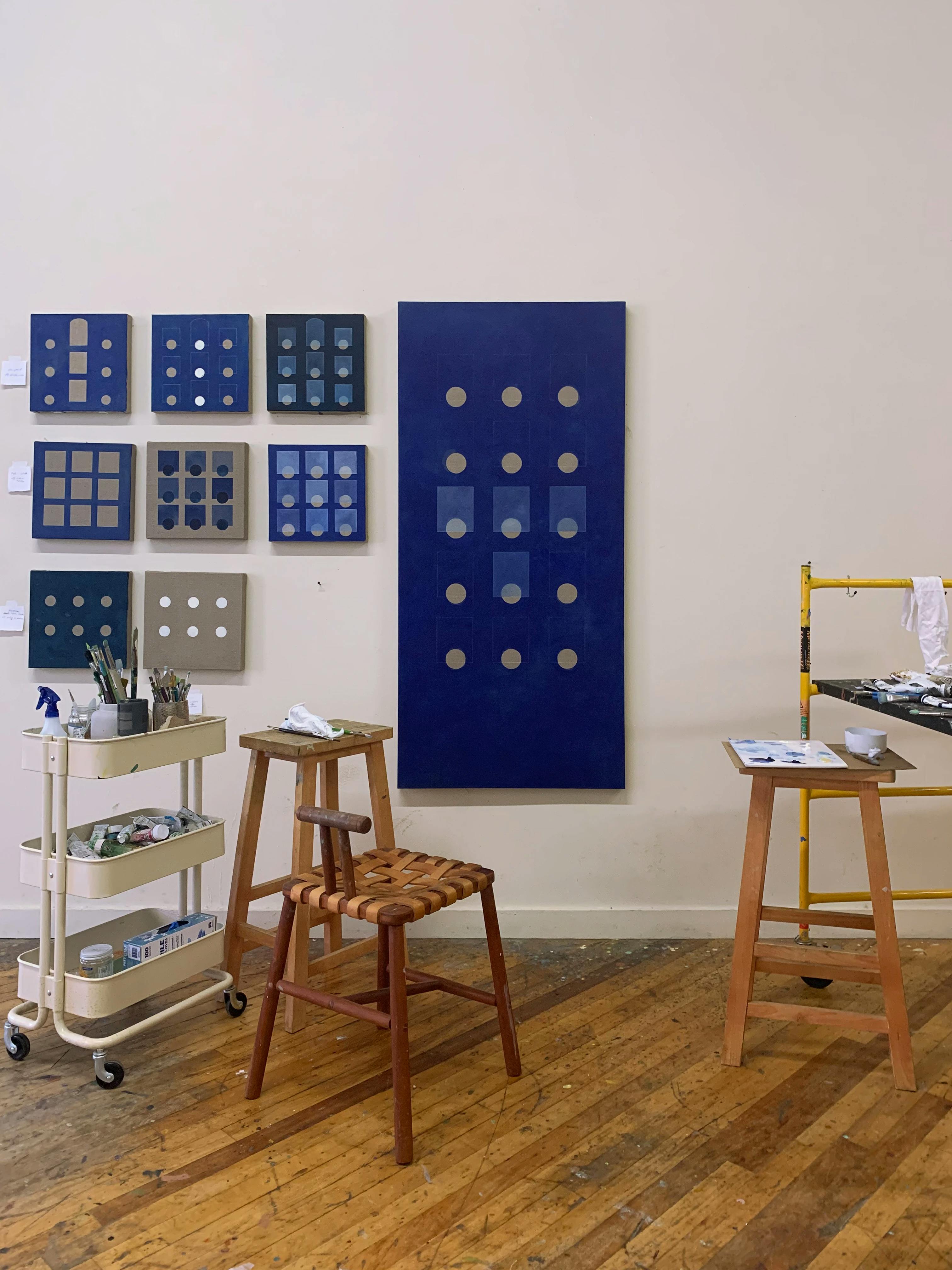 Blue and grey paintings with recurring geometric patterns by artist Carla Weeks installed on a white wall in her studio.