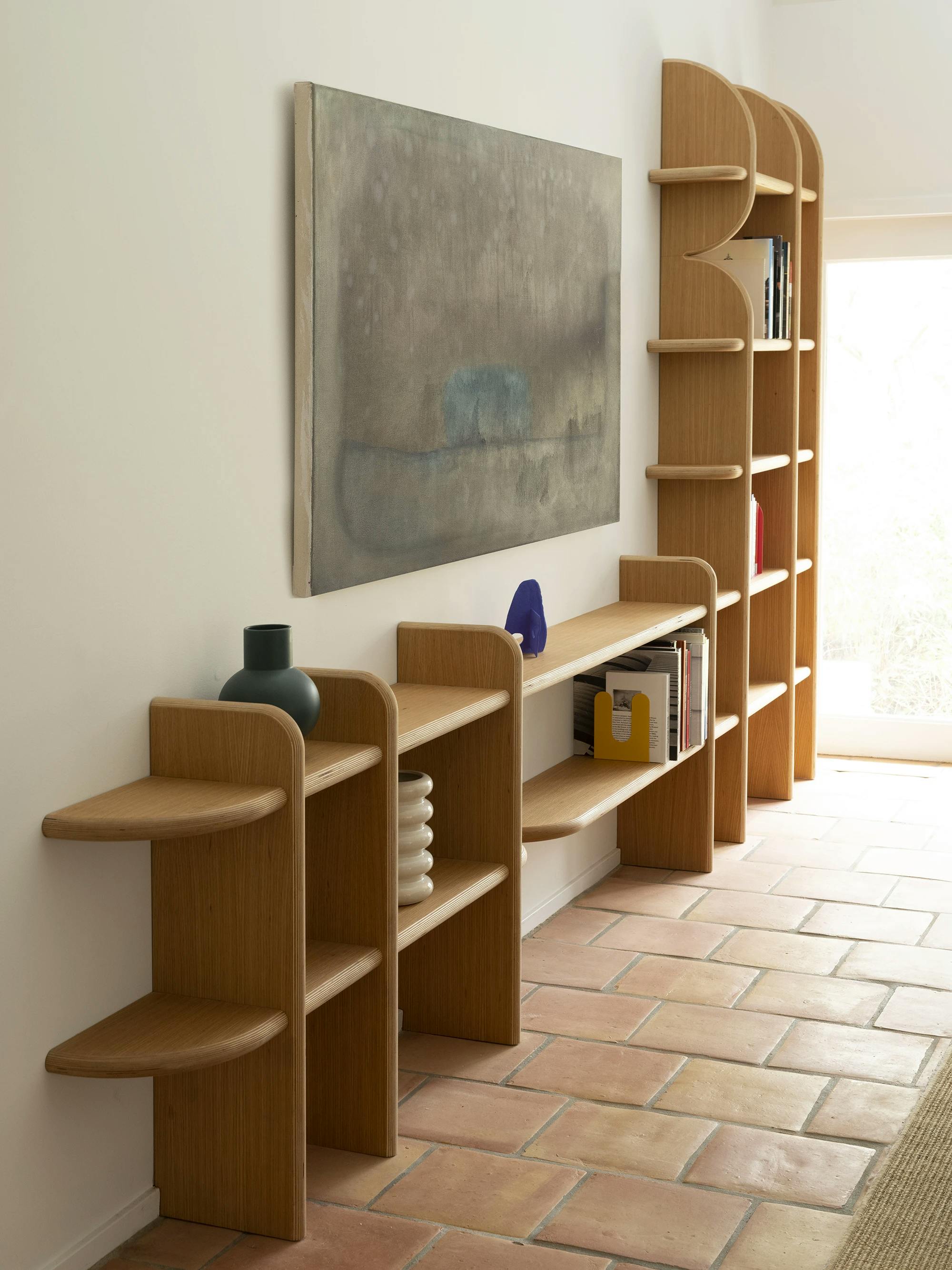 A wooden bookshelf with a painting by Bo Kim hung above. A small blue sculpture by Fitzhugh Karol sits on the bookshelf. 