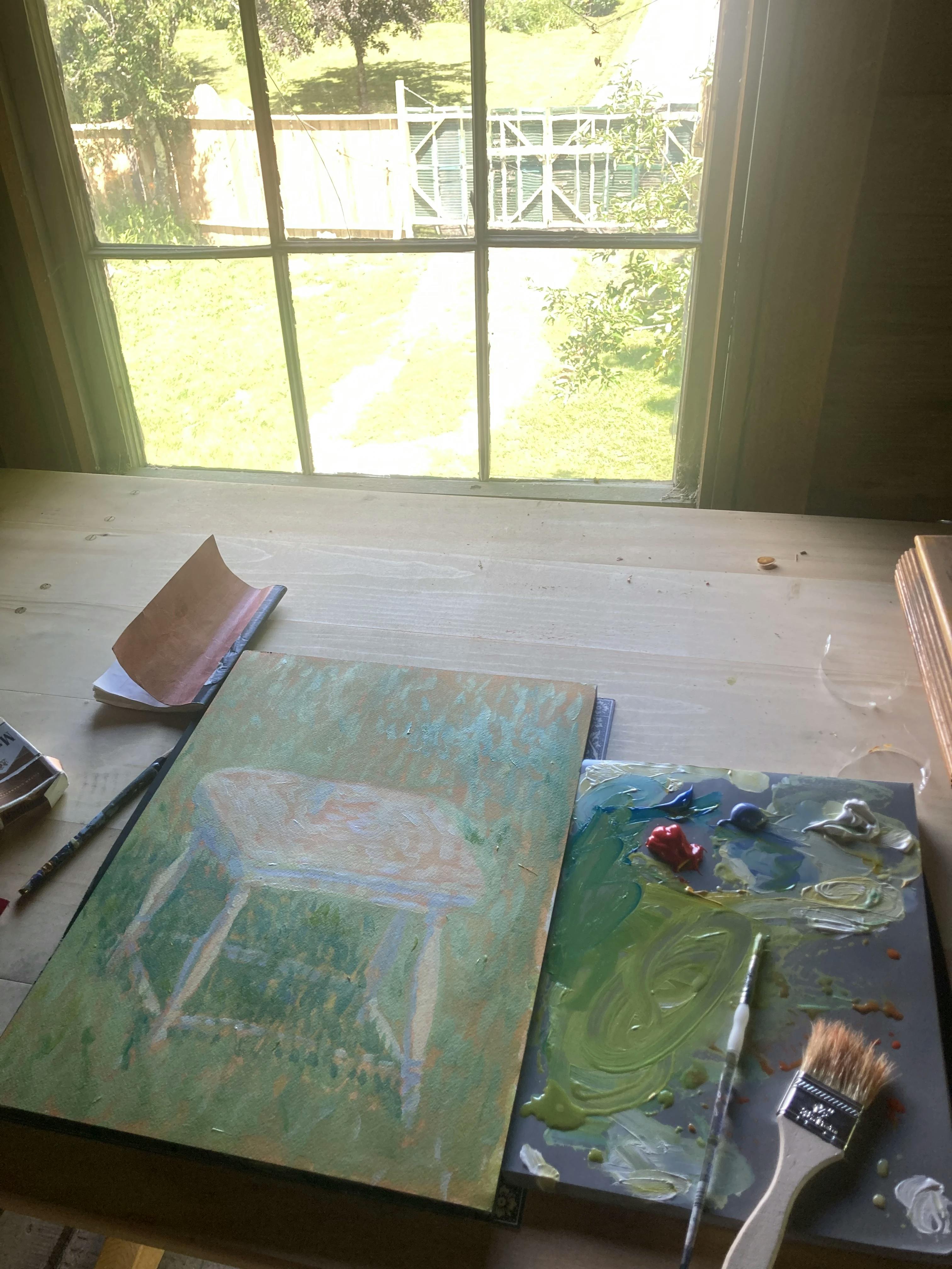 A small painting on paper of a white stool by artist Jackson Joyce on a desk next to a large sunny windowing with a view of a grassy lawn.