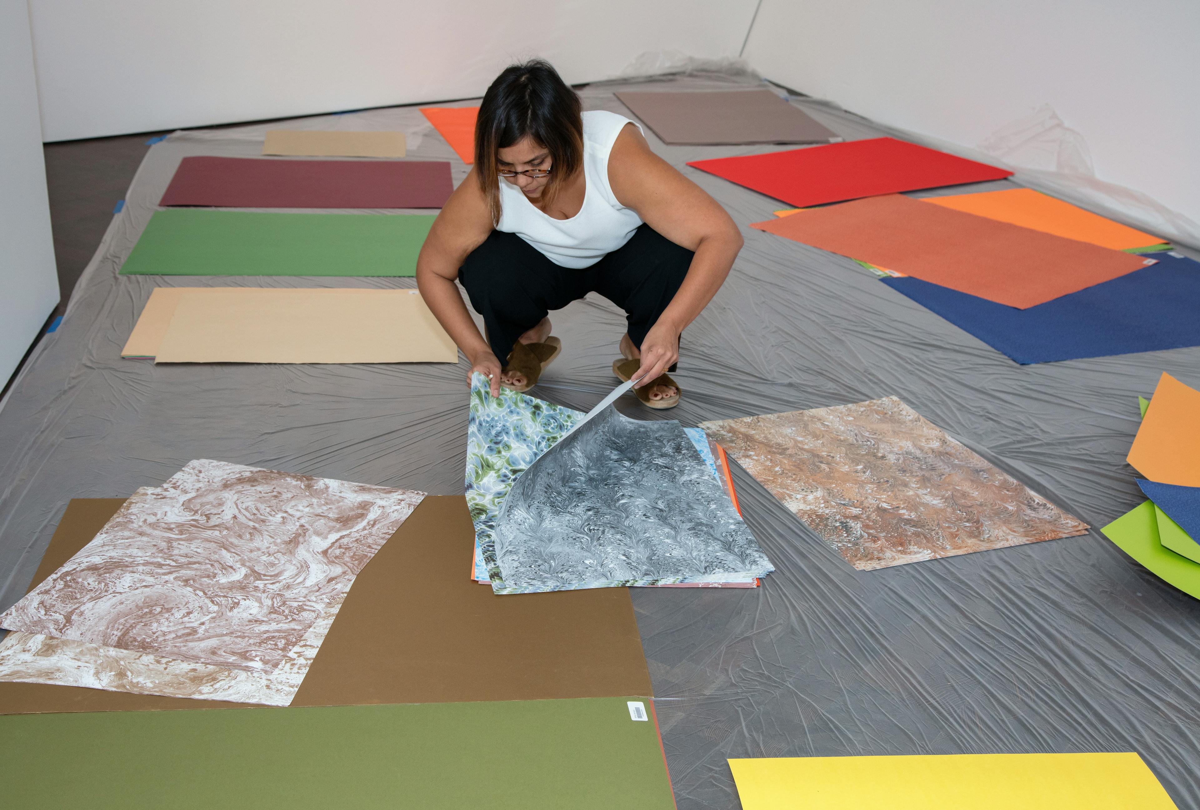 Artist Xochi Solis crouching down on a plastic mat, surrounded by sheets of colorful paper and patterns.