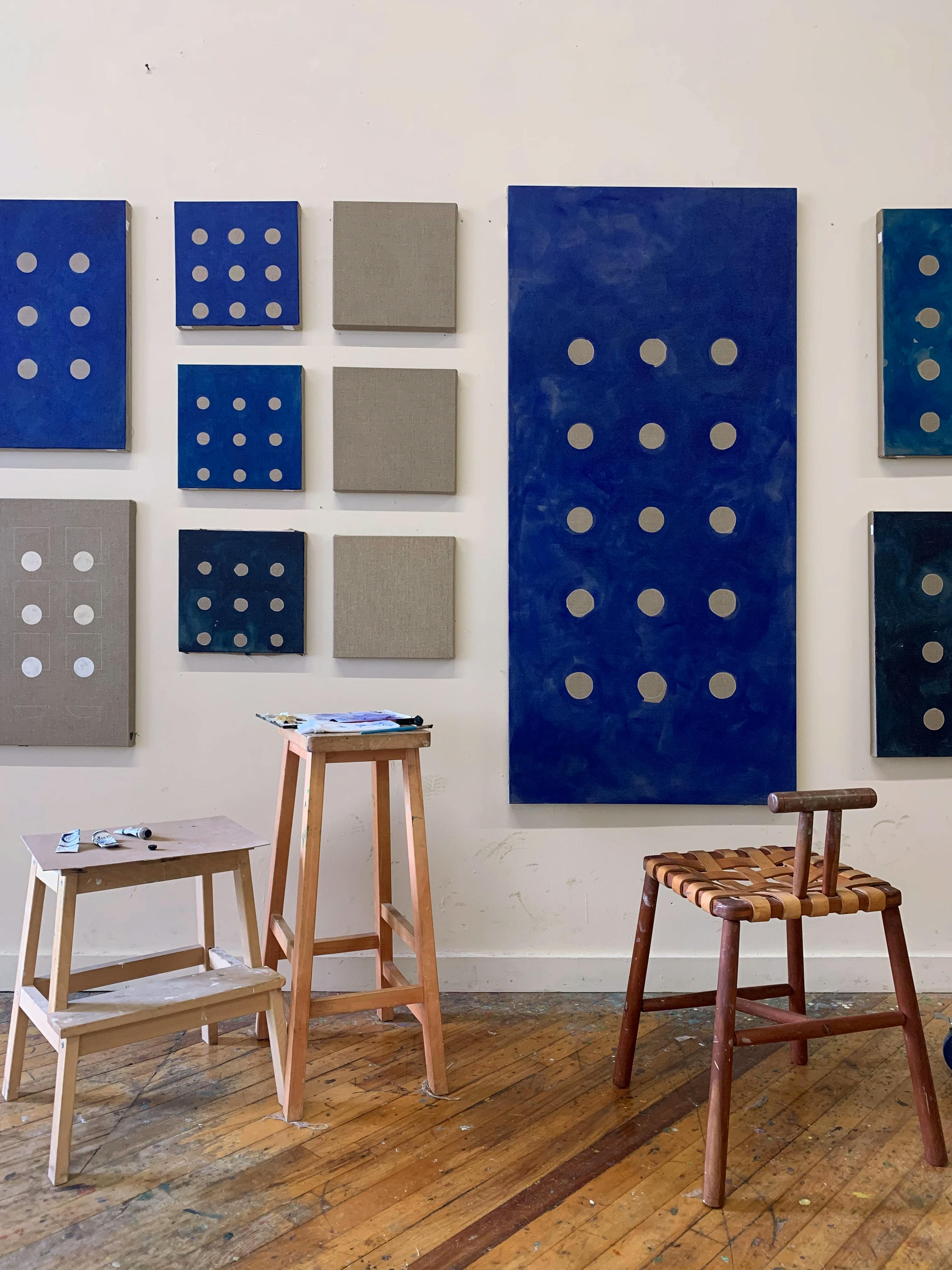 Minimalist, blue and grey oil paintings of different sizes installed on a white wall in artist Carla Weeks' studio.