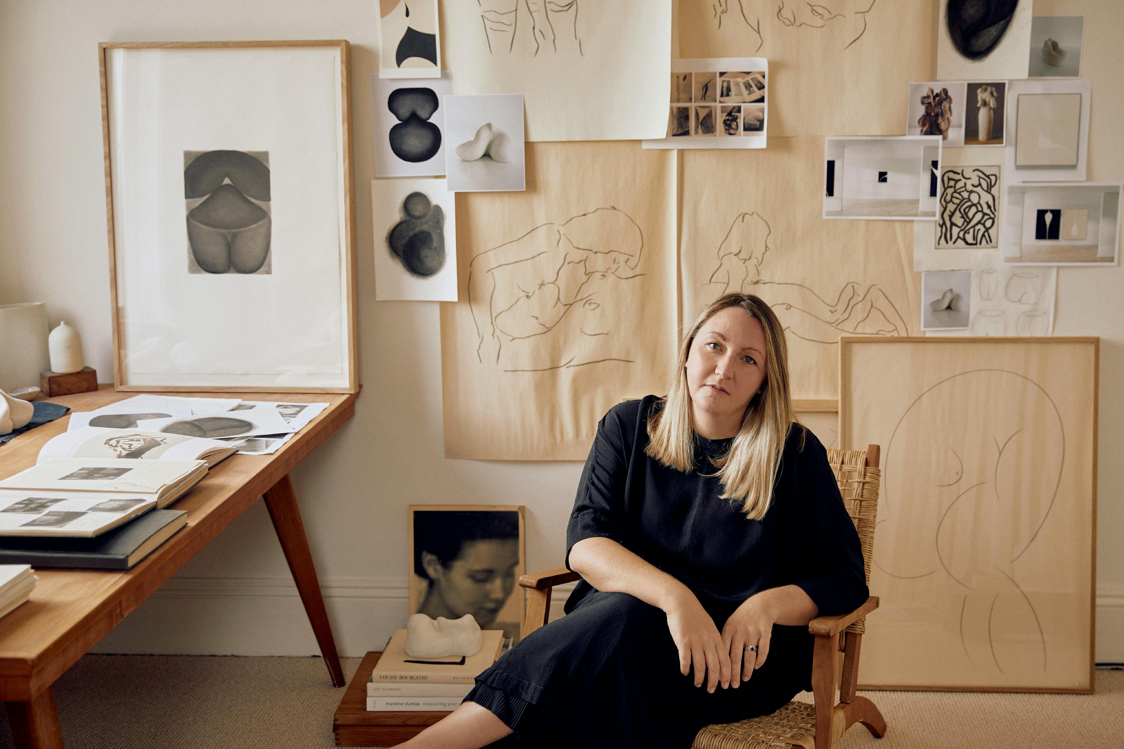 Artist Caroline Walls sits in her studio, surrounded by drawings pinned to the wall and a wood table with books laid out.
