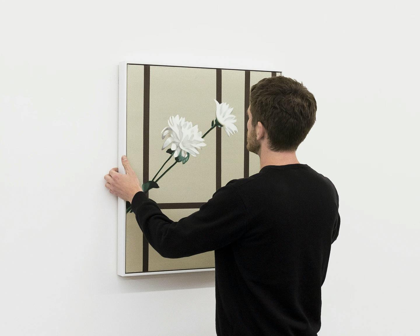 Artist Bryce Anderson wearing a black sweater, holding his painting with Chrysanthemums up against a white wall.