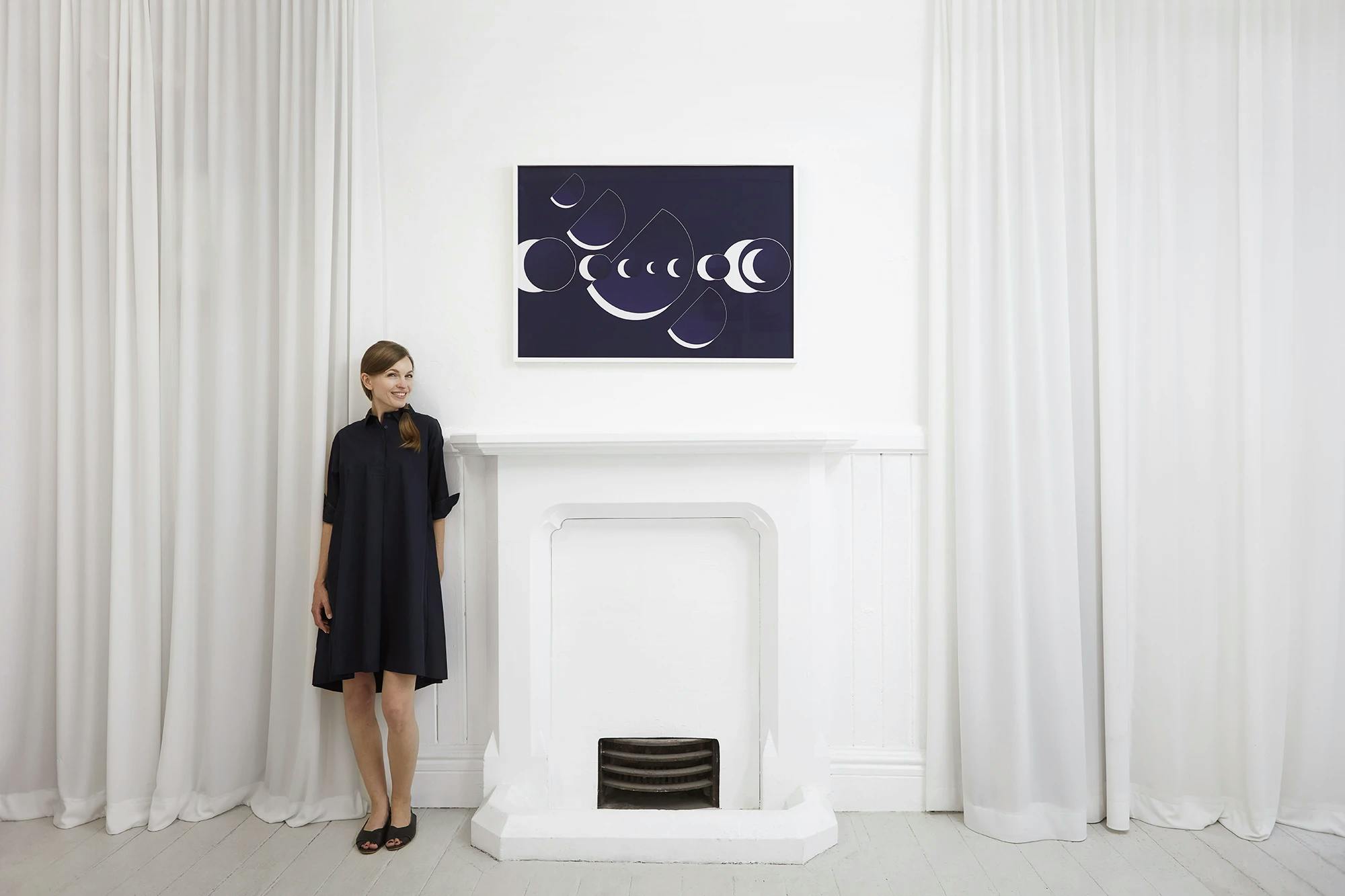 Artist Martina Lang in a navy blue dress standing in a white room next to one of her space prints installed above a white mantle.