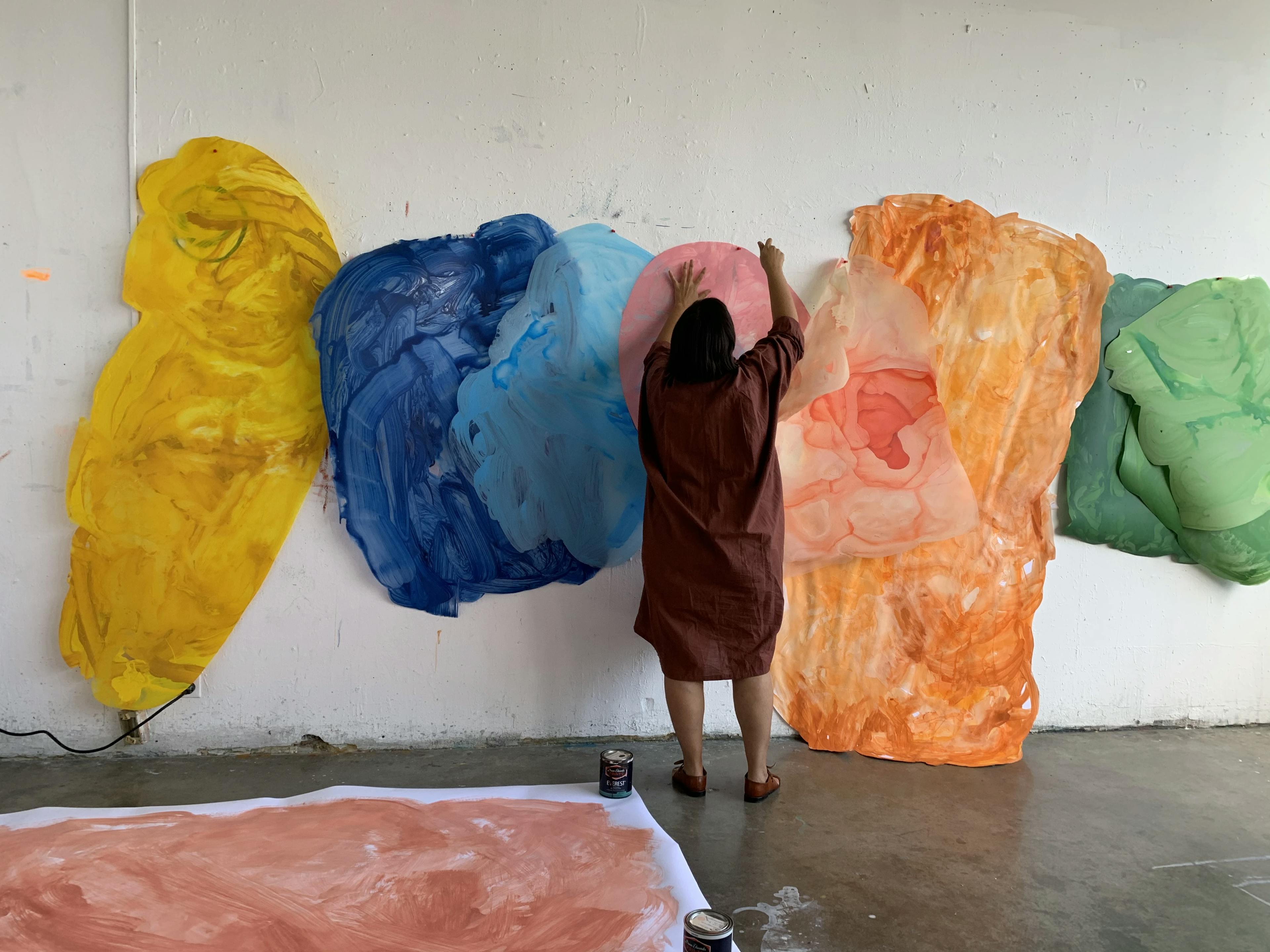 Artist Xochi Solis pinning oversized pieces of brightly colored paper to a white wall in her studio.