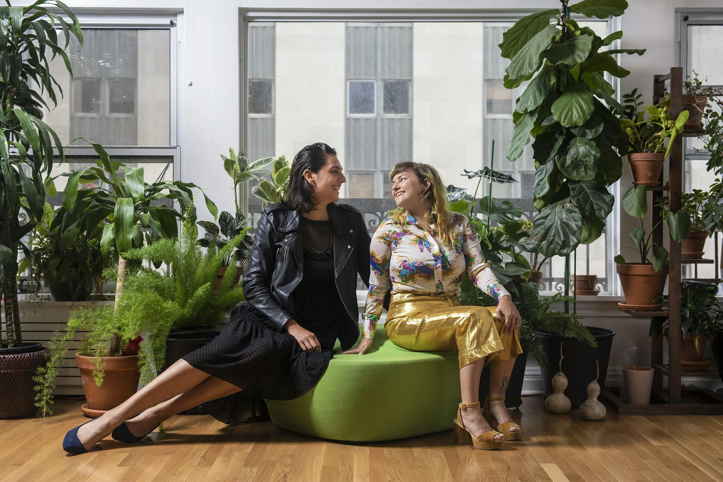 Lauren Hirshfield and Kat Ryals of PARADICE PALASE sitting side-by-side on a green chair, surrounded by multiple potted plants in an office with a large window.