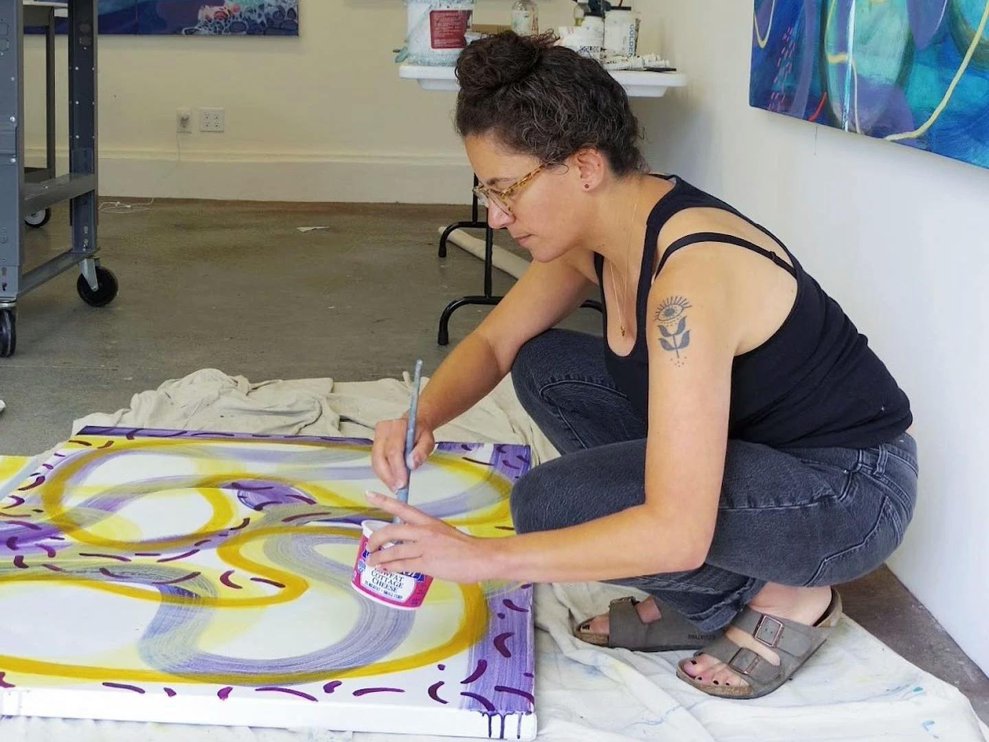 Artist Aliza Cohen crouching down in her studio and painting abstract yellow and purple lines on a canvas.