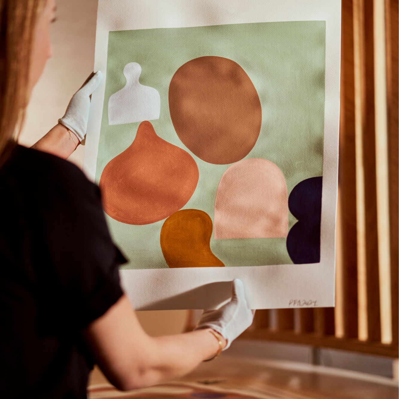 Woman in black shirt with white gloves holds an abstract artwork above a table.