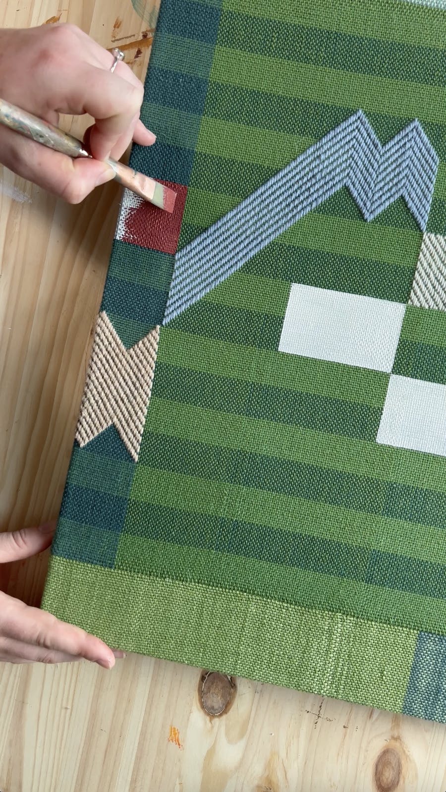 An aerial view of artist Sarah Sullivan Sherod painting a red line on a green striped, handwoven artwork.