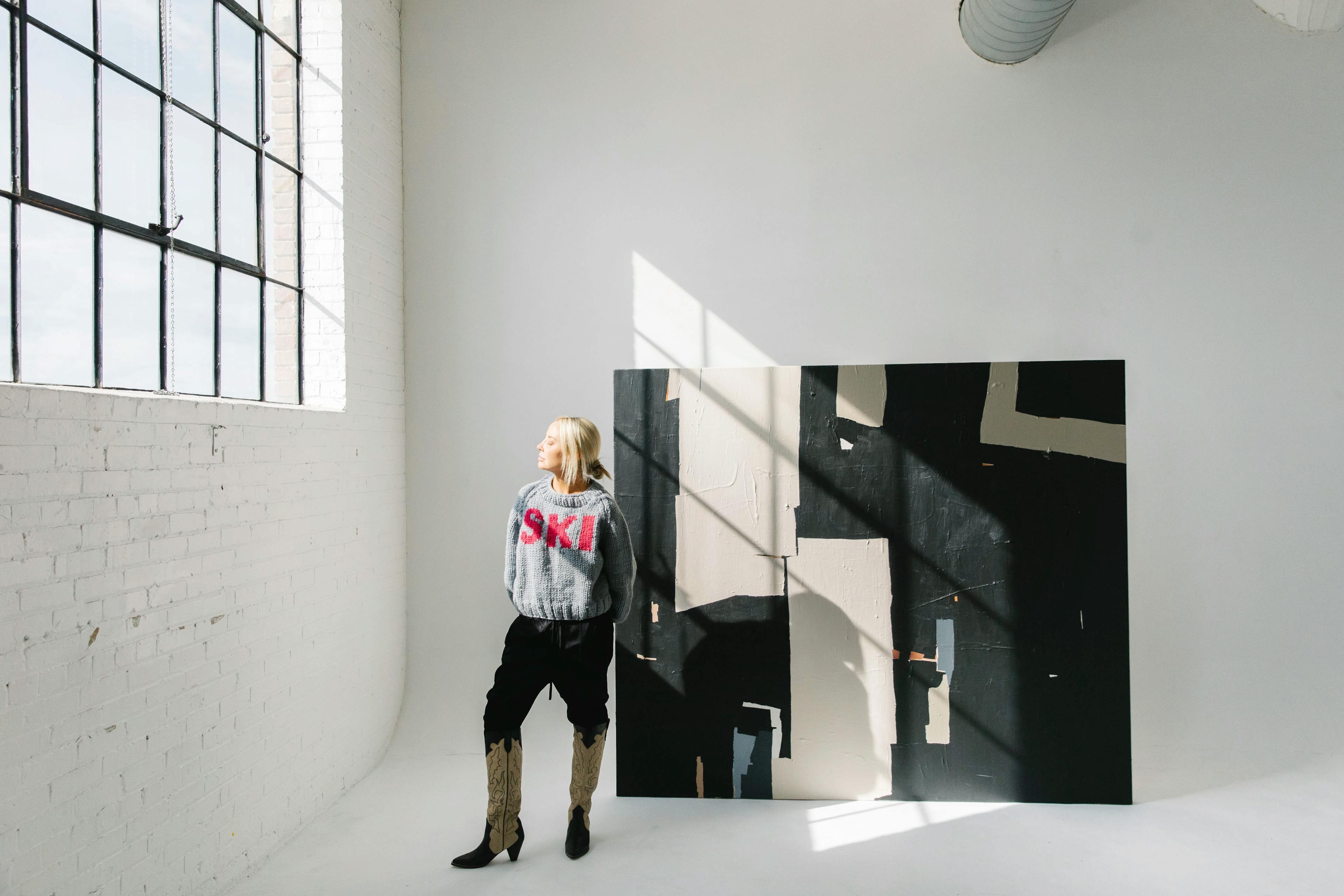 Artist Holly Addi standing in her white-walled studio next to an oversized, abstract geometric painting.