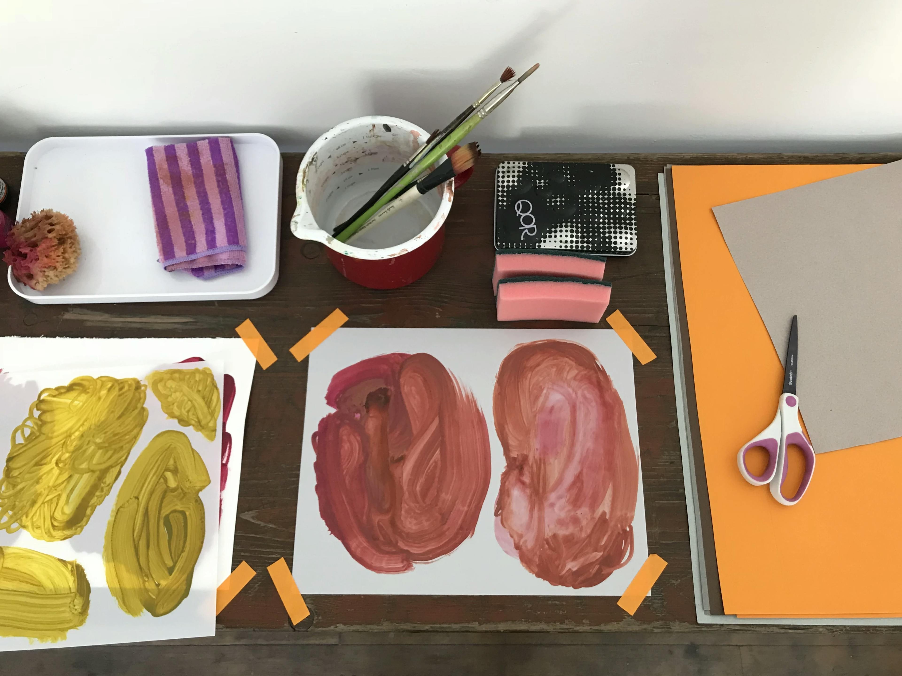 Two in-progress works by artist Xochi Solis with swaths of colors, one yellow and one red, taped to a wooden surface in her studio.