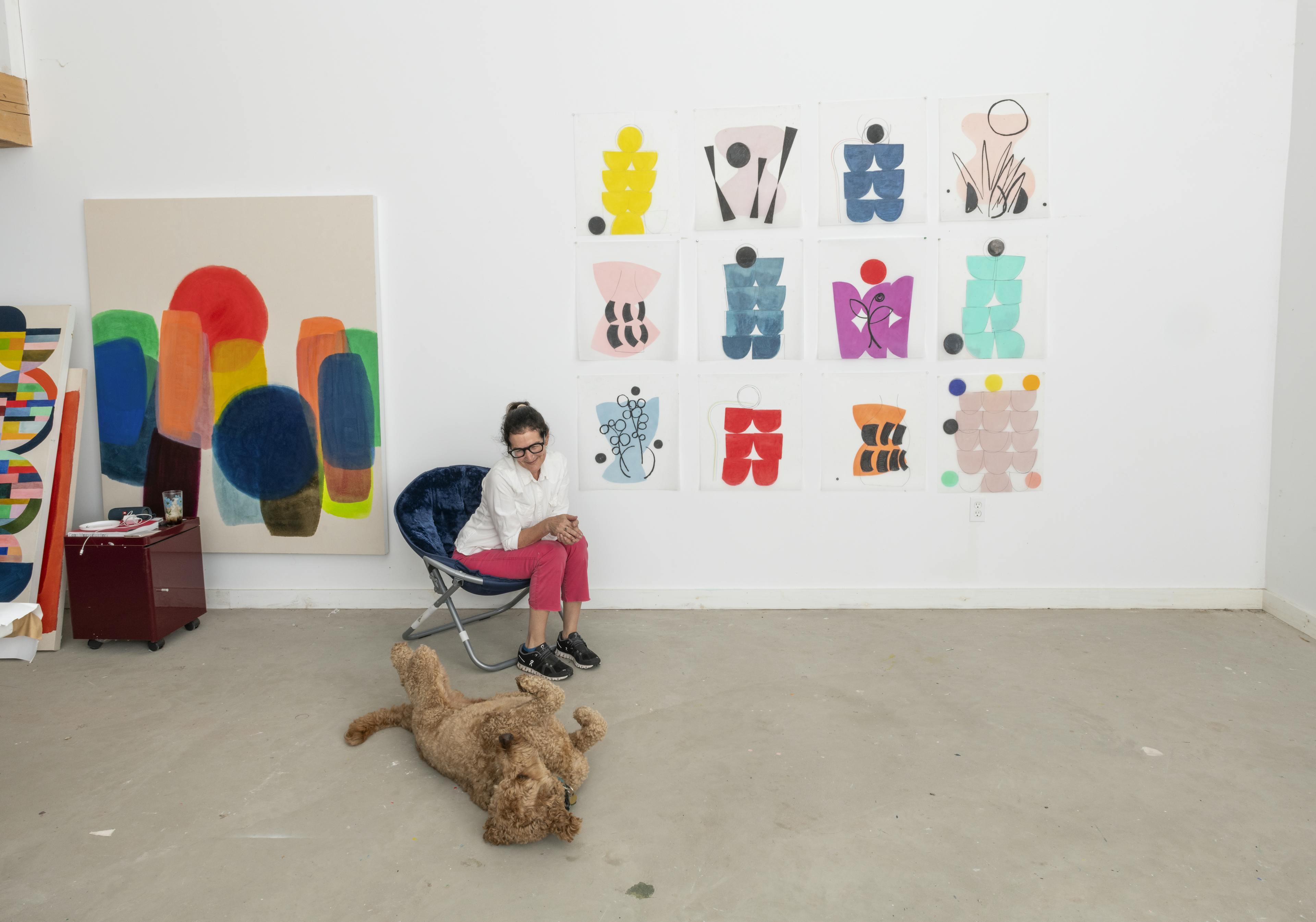 Artist Vicki Sher sitting in a blue chair in her studio surrounded by her drawings, looking at her dog rolling on the floor.