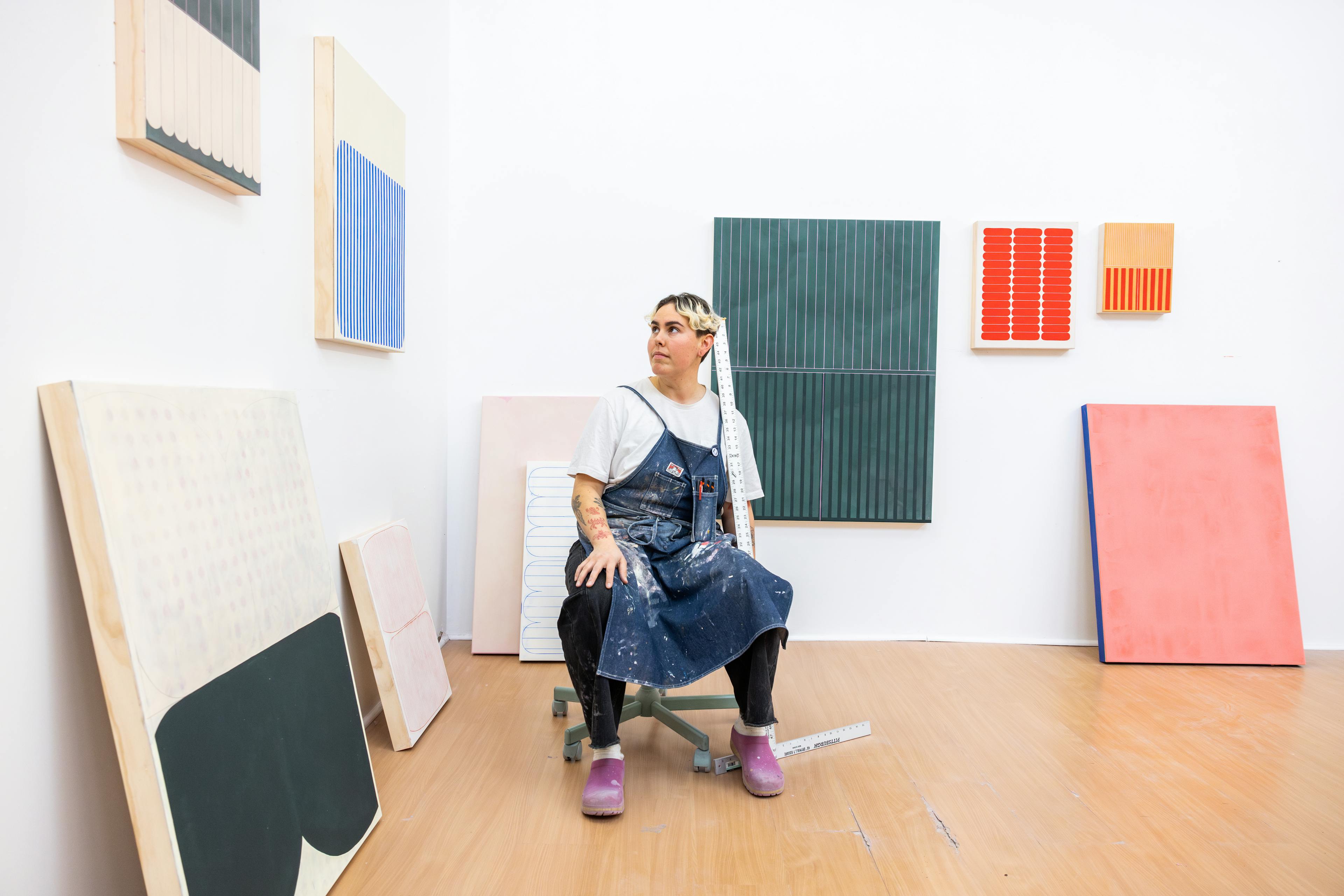 Artist Arielle Zamora in the studio sitting on a stool surrounded by their minimalist, abstract paintings.