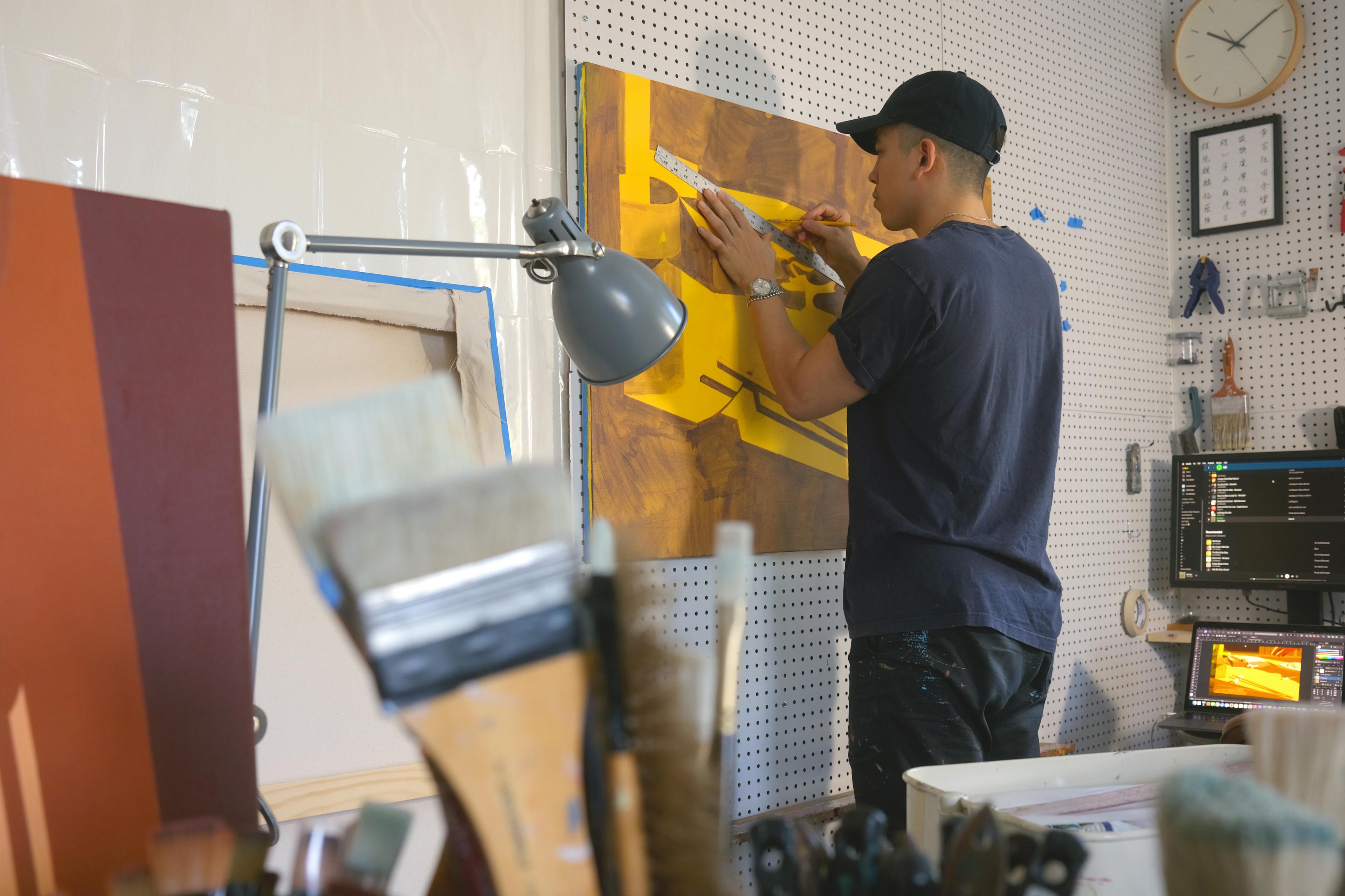 Artist Adrian Kay Wong in his studio using a ruler to create a straight line on a yellow canvas.