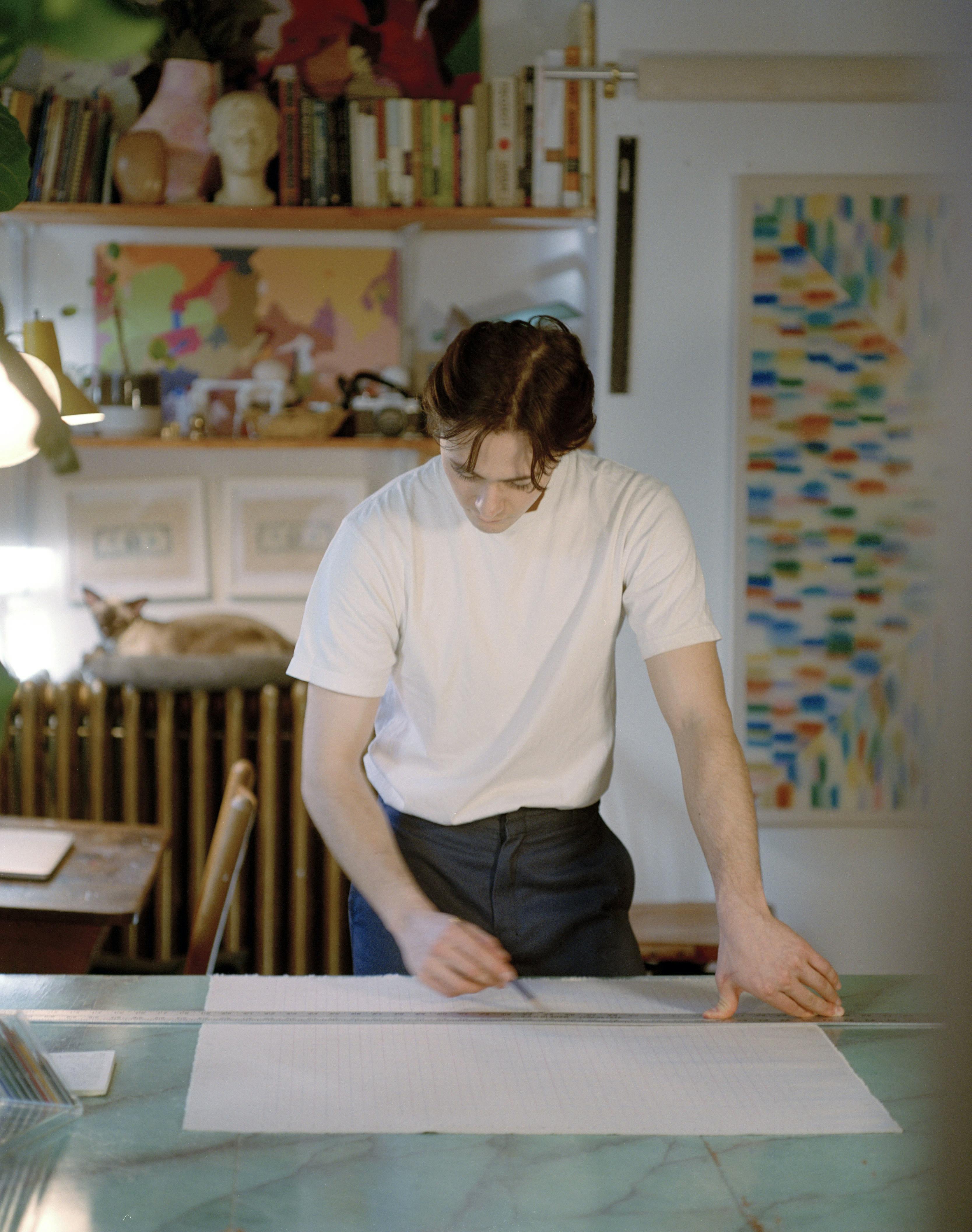 Artist Devon Reina in his studio using a ruler to draw straight pencil lines on paper.