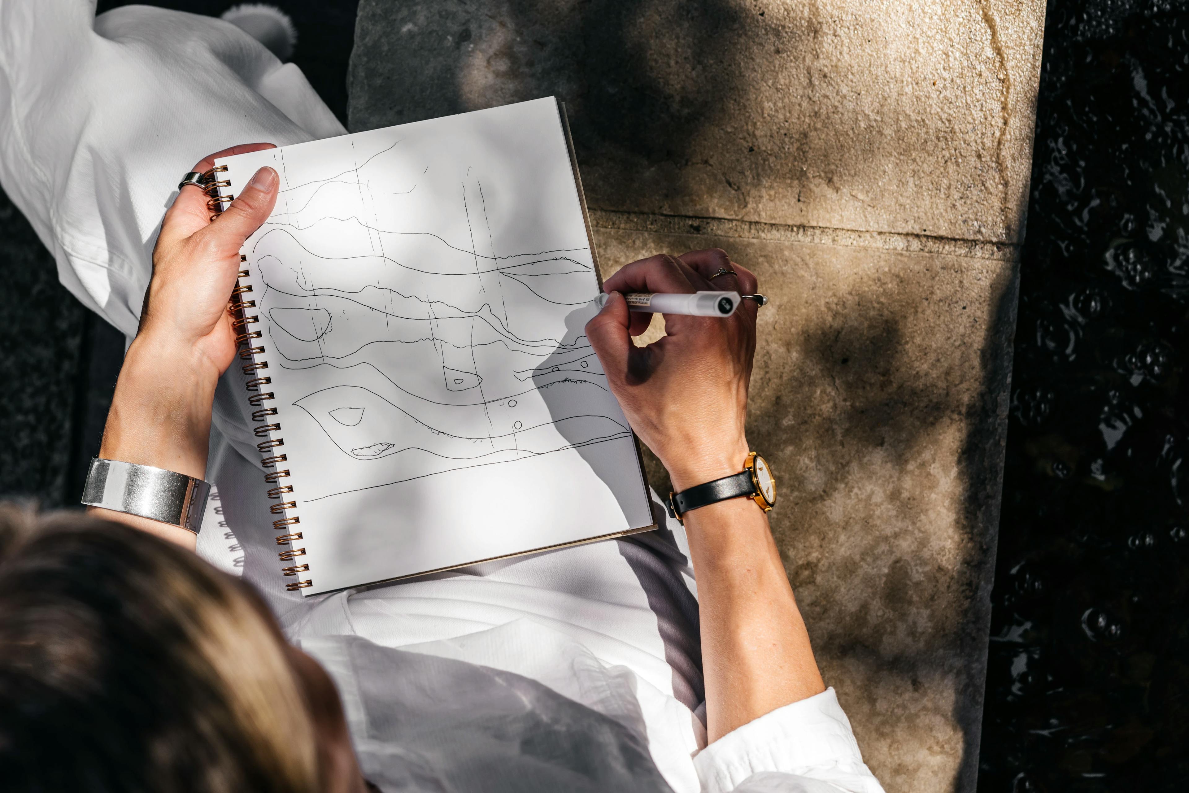 An overhead look at Laura Naples sketching curved lines in a notebook while sitting on the ground at MacArthur Place.