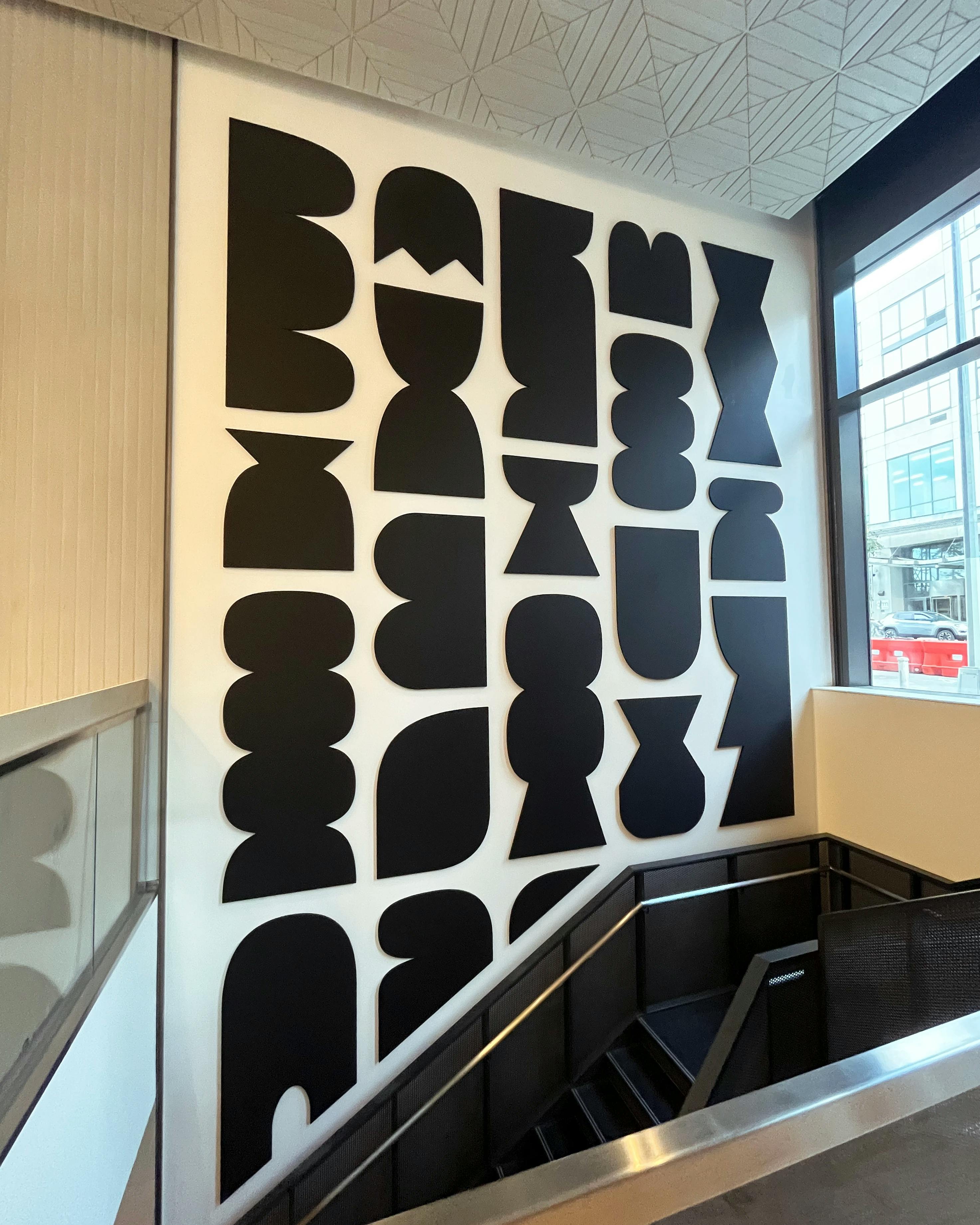 A custom floor-to-ceiling mural of black geometric shapes on a white wall by artist Dan Covert in the stairwell of a modern residential building.
