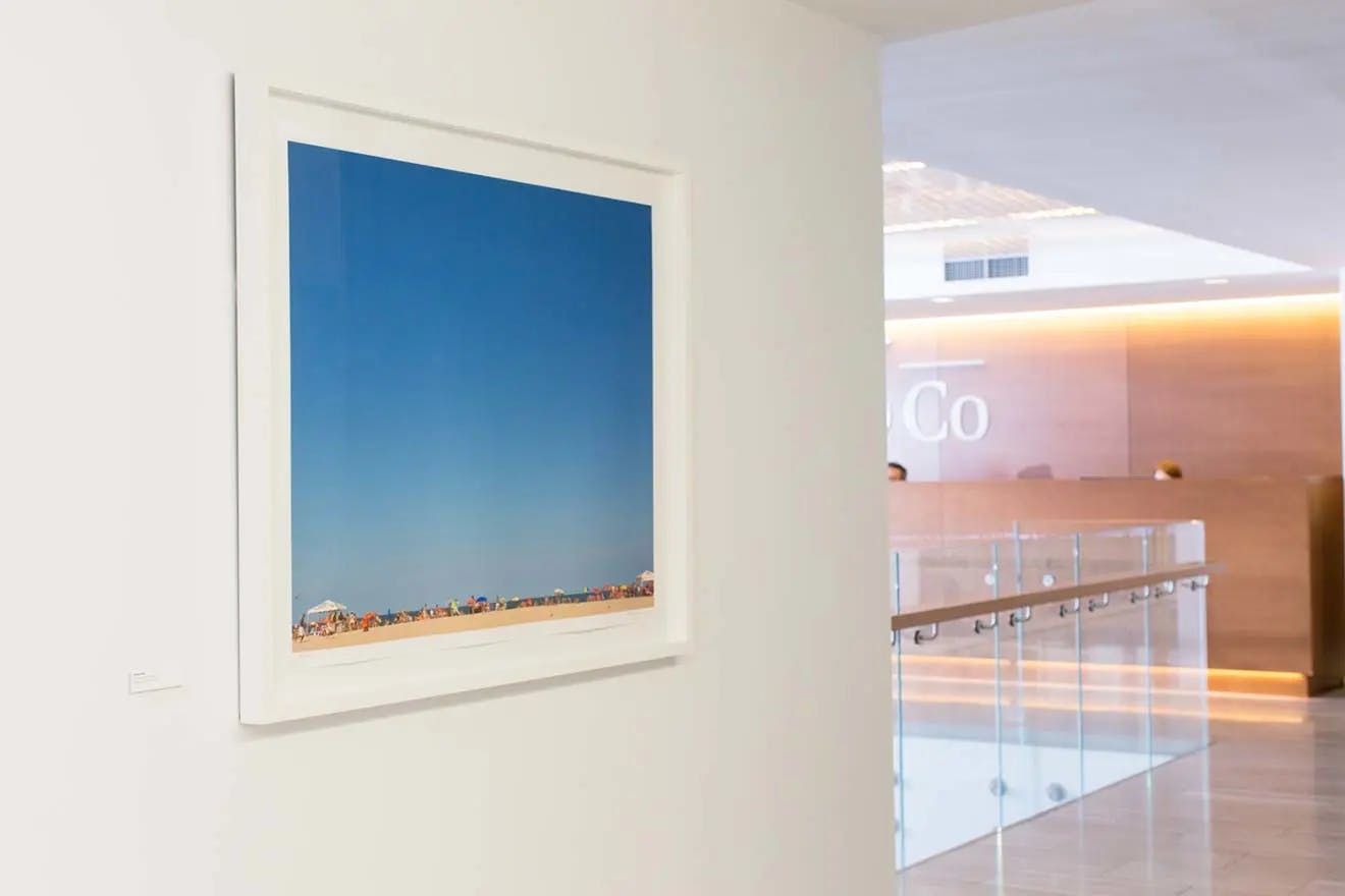 A framed photograph of a beach with clear blue skies by artist Ashok Sinha installed on a white wall in a modern office building.