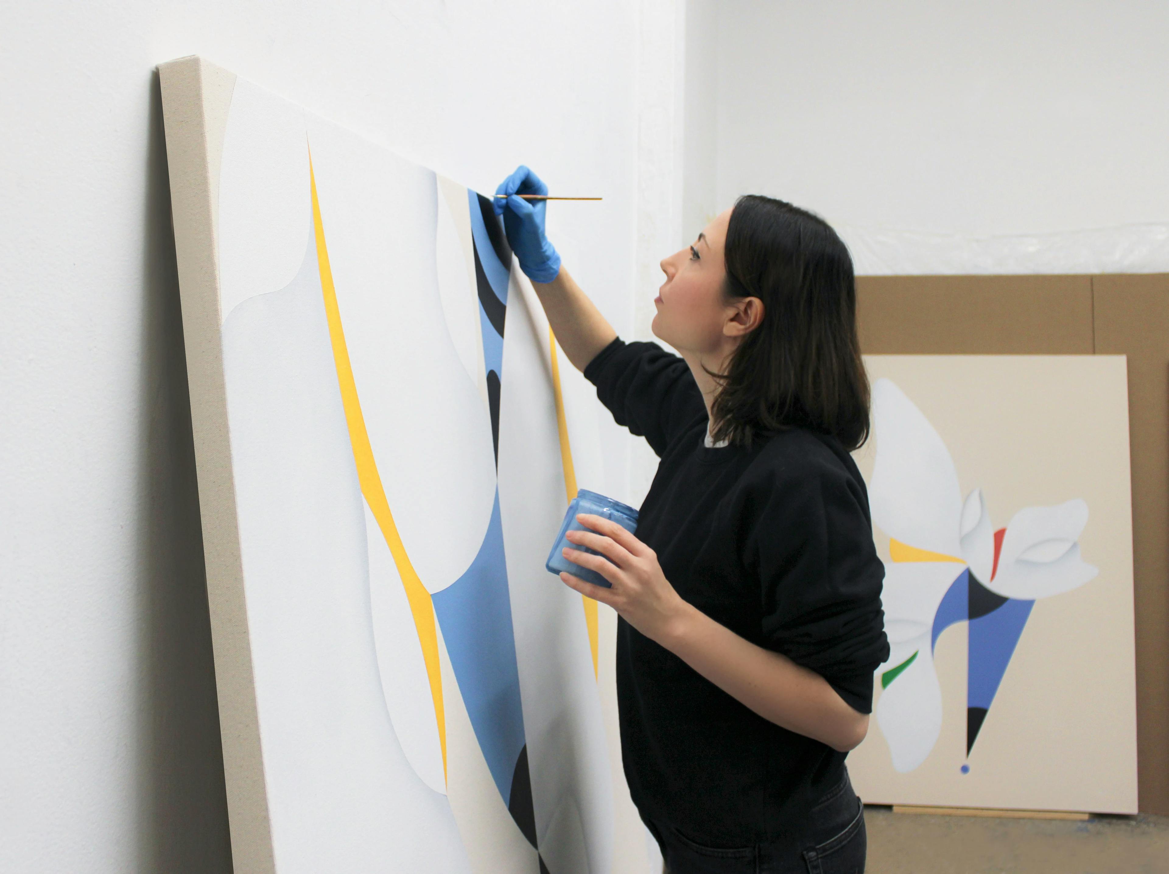 Artist Senem Oezdogan holding a small can of light blue paint and painting a large canvas in her studio.