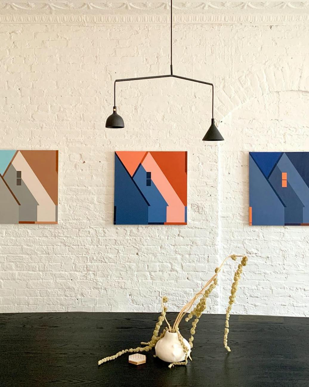 Three abstract paintings of rooftops hang in a line on a white brick wall over a black table at Uprise Art.