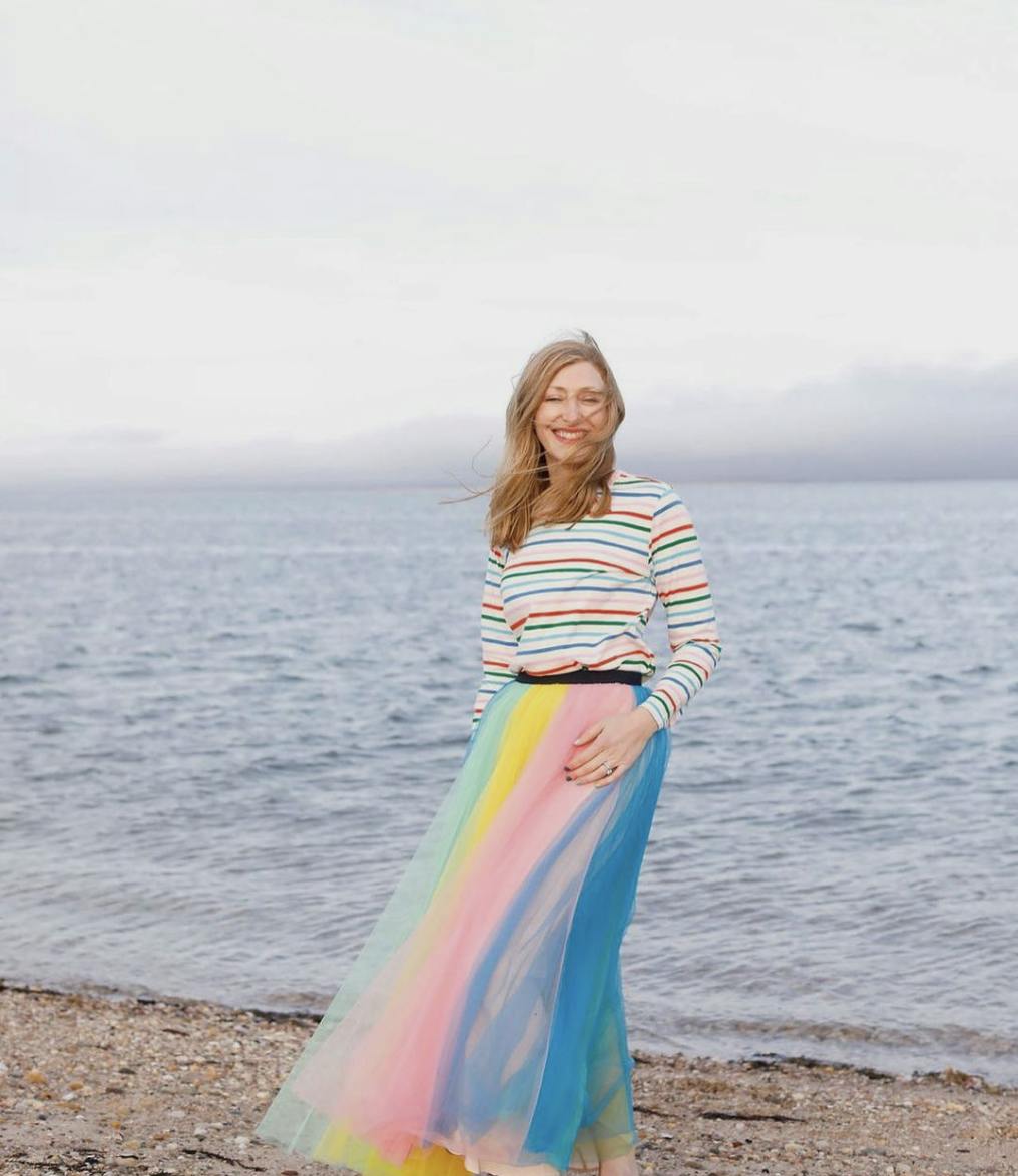 Ingrid Fetell Lee standing in front of an ocean wearing a striped shirt and a long, pastel-colored skirt blowing in the wind.