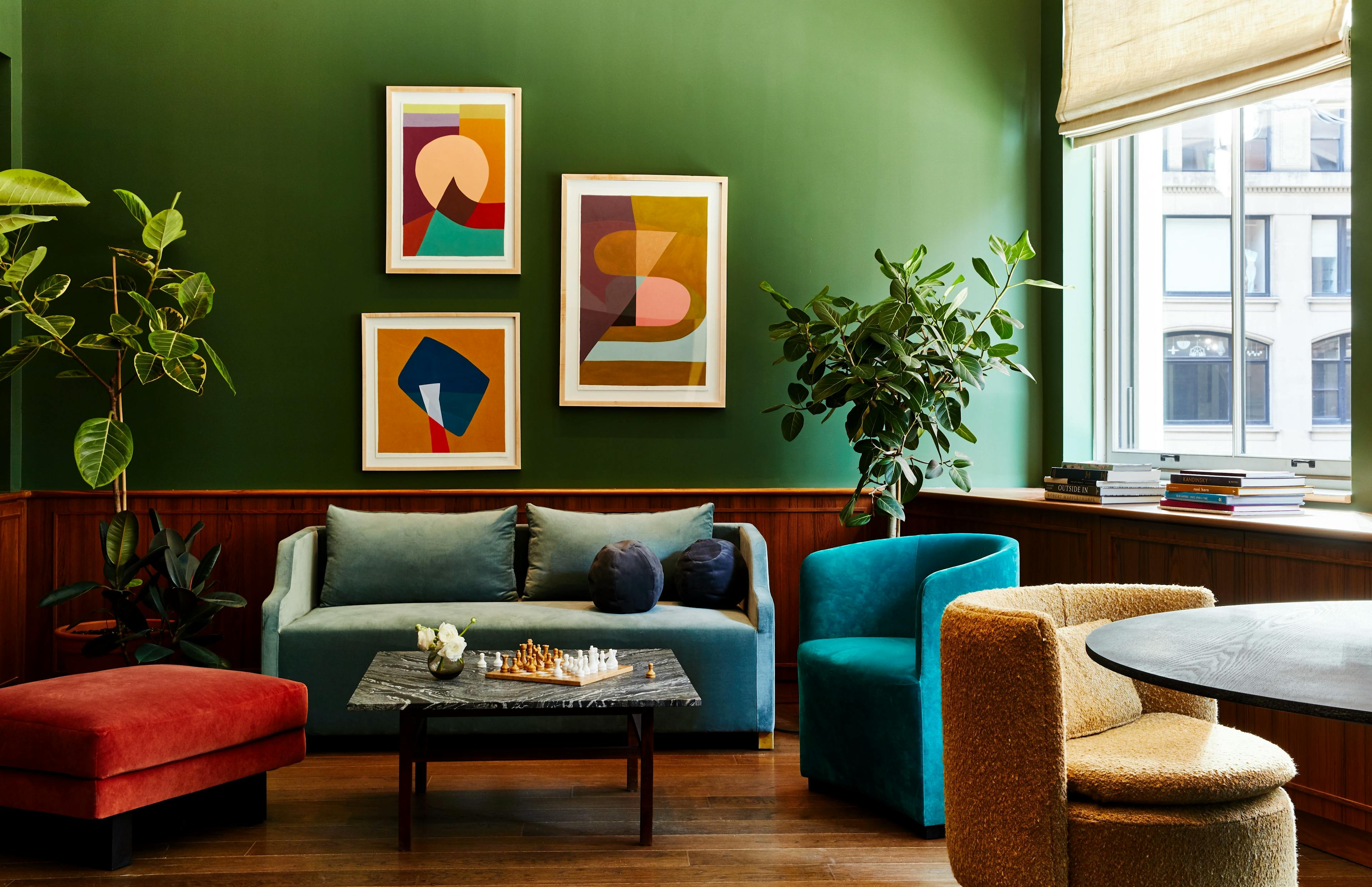 Three framed geometric paintings by Kristin Texeira on a dark green wall in a lounge area with a sofa and armchairs at the Chief Flatiron clubhouse.