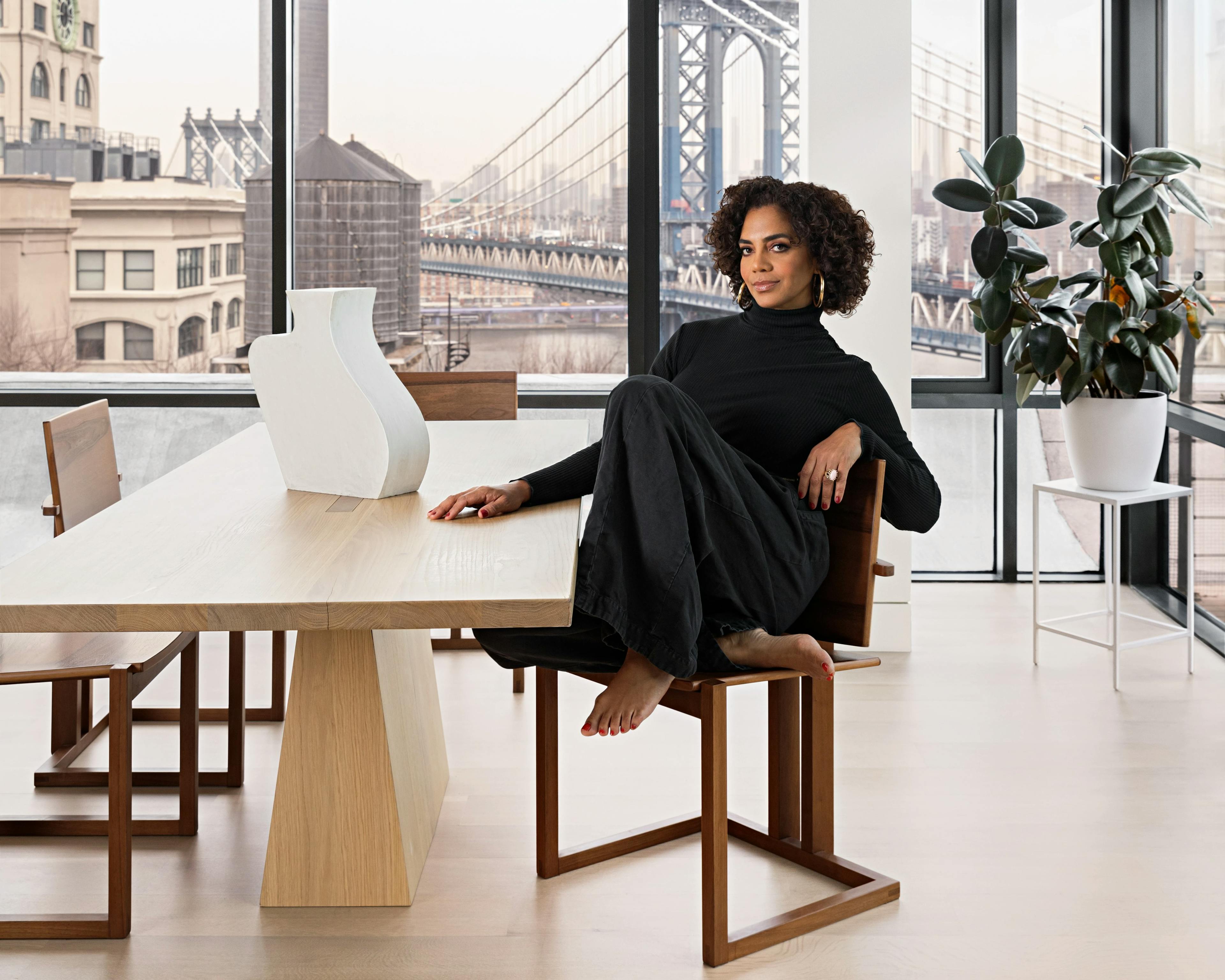 Designer Danielle Colding wearing a black turtleneck and pants, sitting at a wooden table with a white sculpture by artist Dan Covert on top.