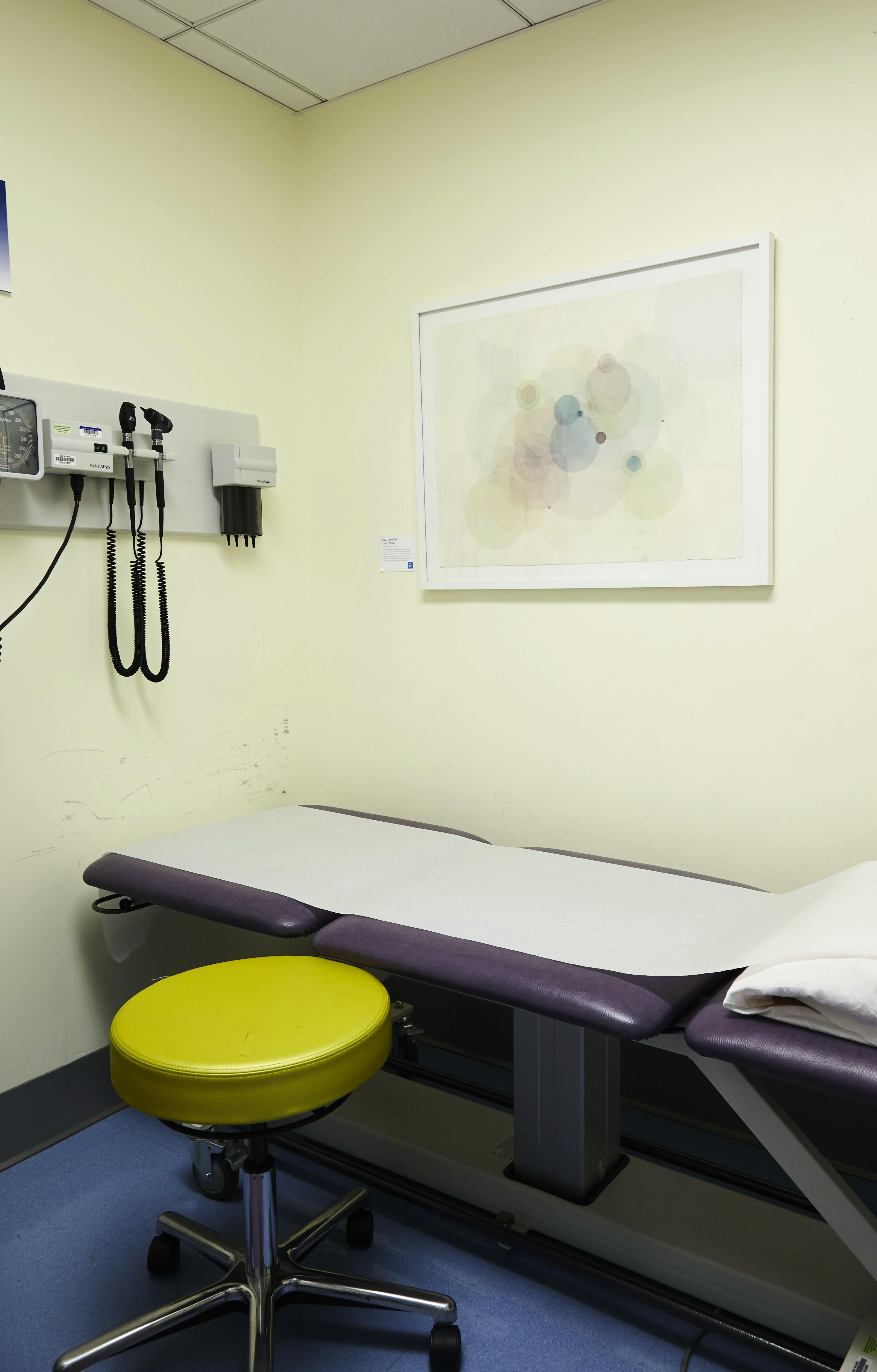 Framed artwork with layered, concentric circles by artist Evan Venegas installed on a beige wall above a purple exam table.