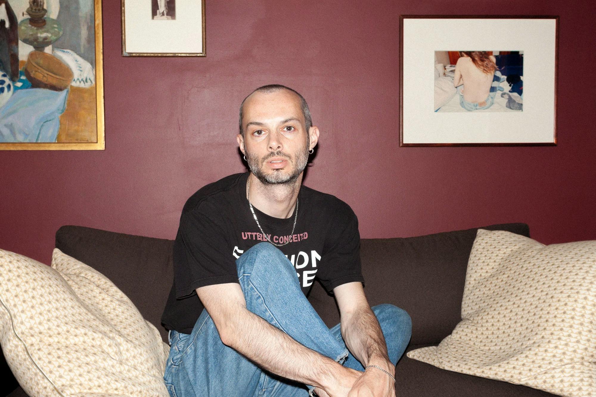 Portrait of Sean Santiago wearing a brown graphic tee and blue jeans, sitting on a sofa in front of a maroon-colored wall with framed artwork.