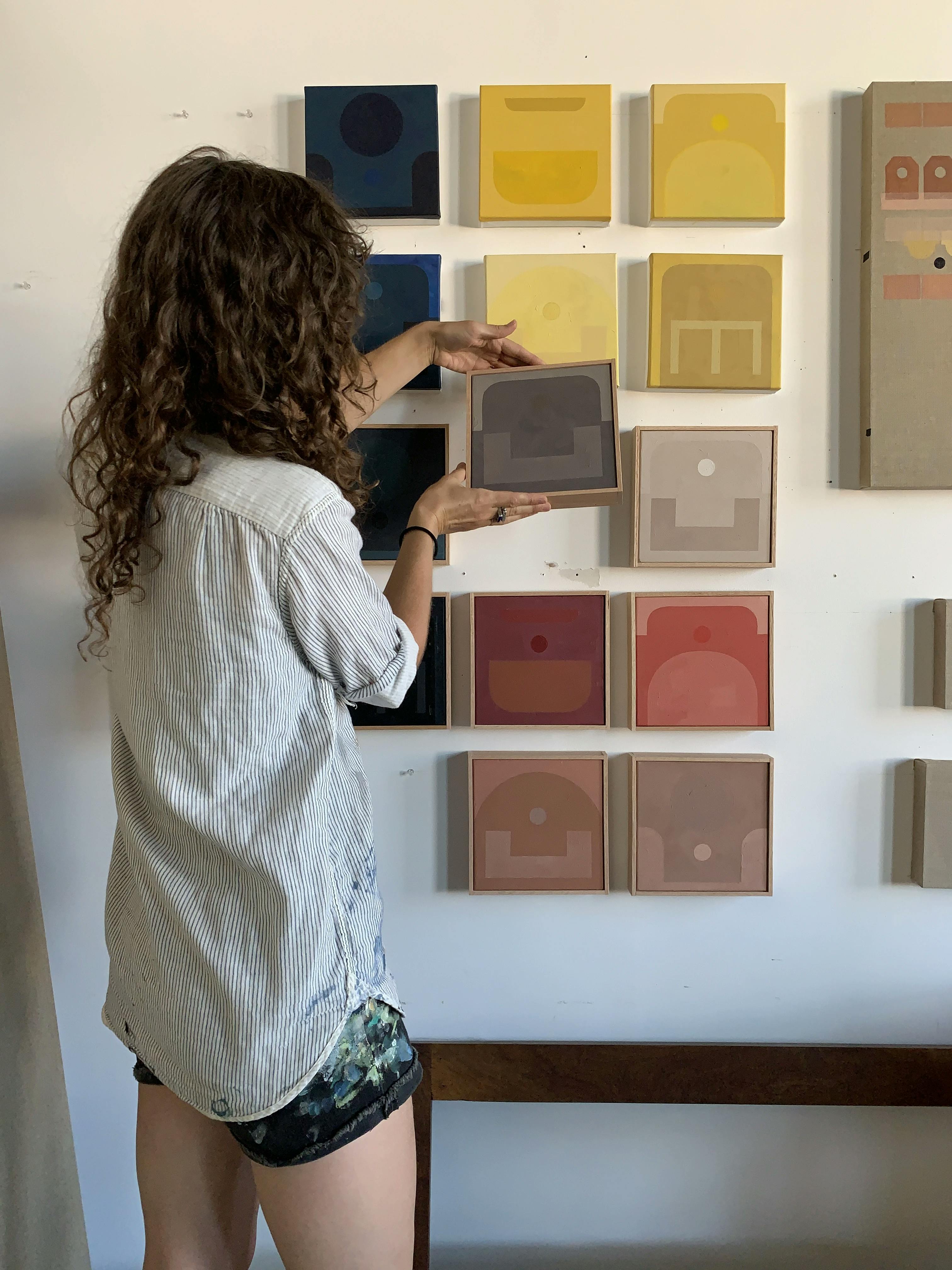 Artist Carla Weeks inside her studio in front of a grid of her small monochrome paintings.