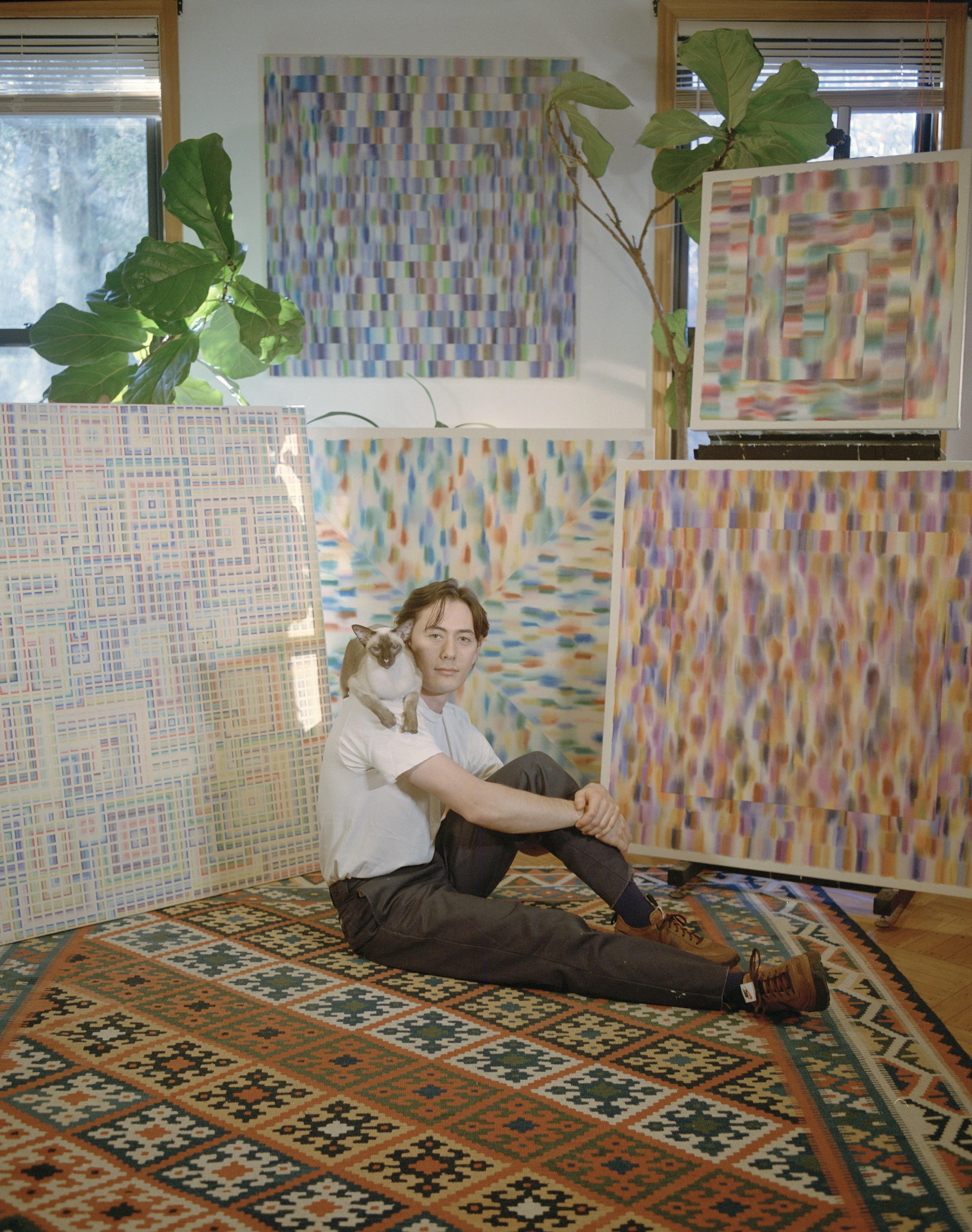Artist Devon Reina sitting on a carpet with a cat on his shoulder surrounded by his paintings.