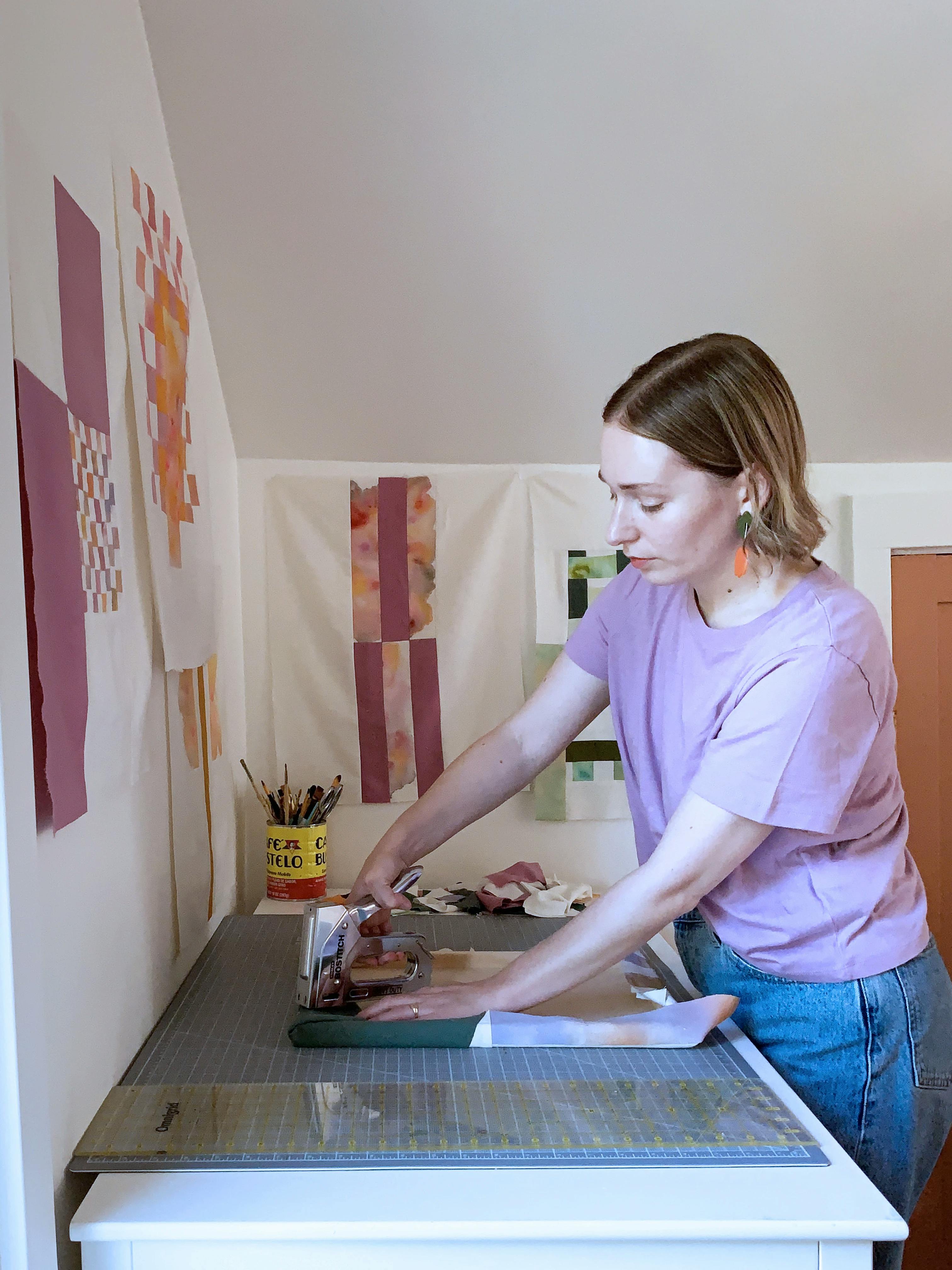 Artist Anastasia Greer wearing a purple shirt in her studio using a staple gun on her stitched silk work.