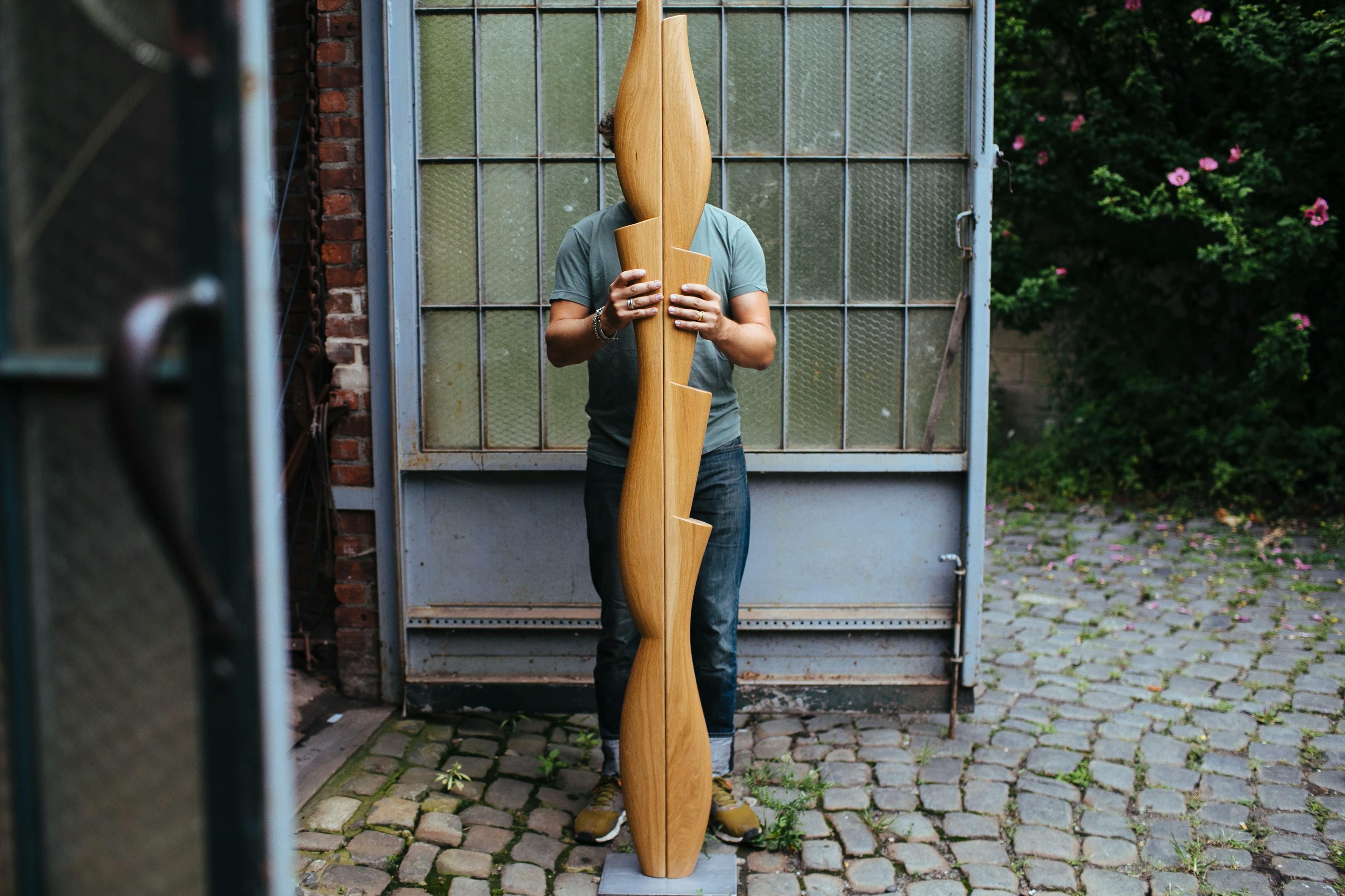 Artist Fitzhugh Karol standing outside his studio on cobblestone pavers holding a tall wooden sculpture.