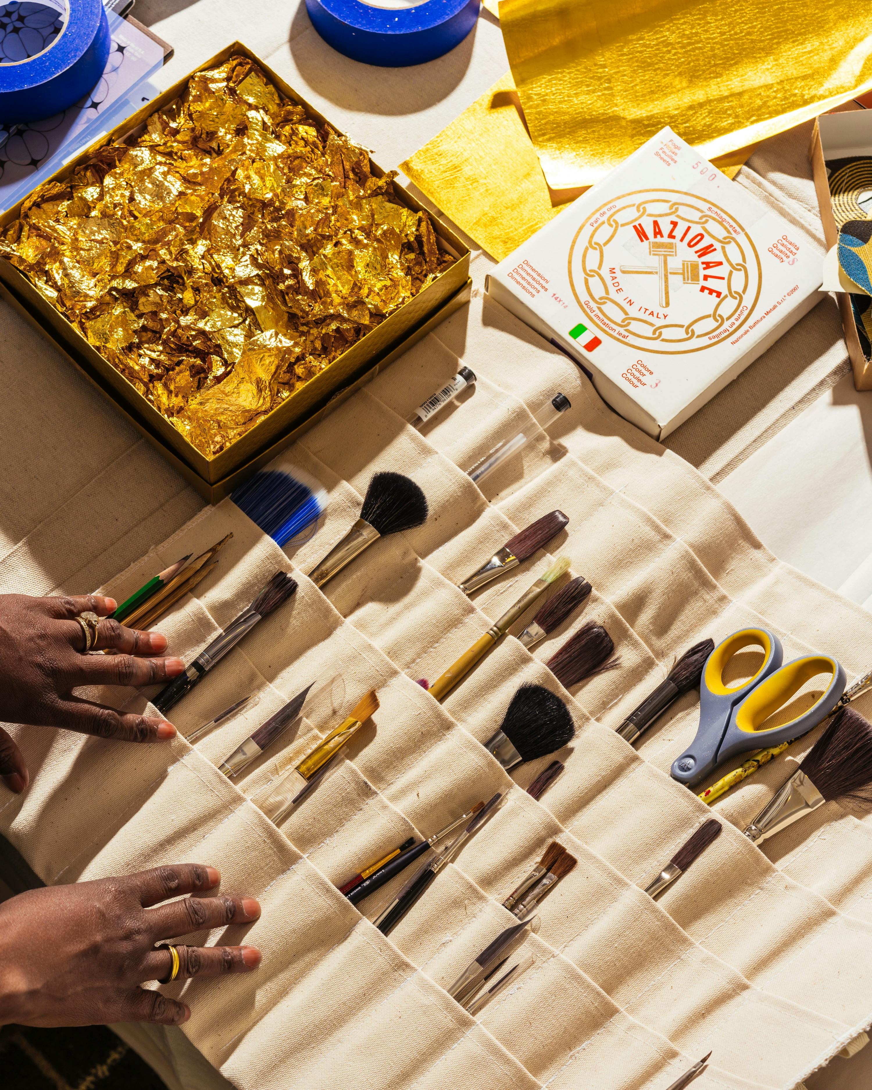 Artist Lisa Hunt with a box filled with gold leaf next to multiple paintbrushes.