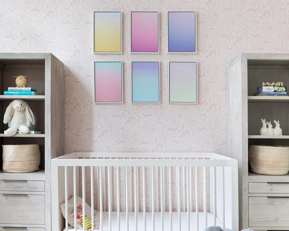 Six framed photographs of pastel-colored, gradient skies by artist Jordan Sullivan on a light pink wall between two bookshelves and above a white crib.