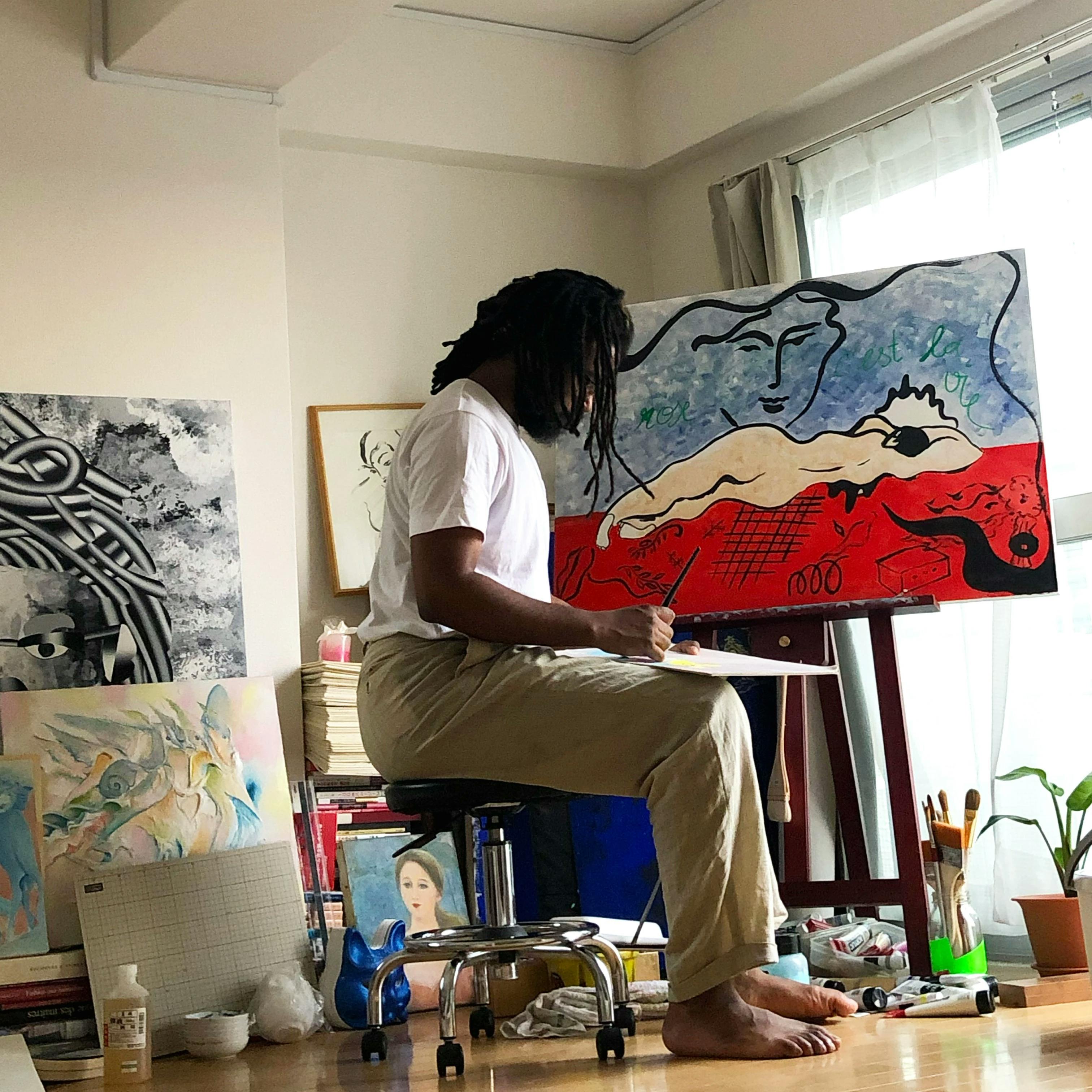 Artist Ba Ousmane in his studio, sitting on a stool and working on a red, white, and blue painting of a reclining woman.