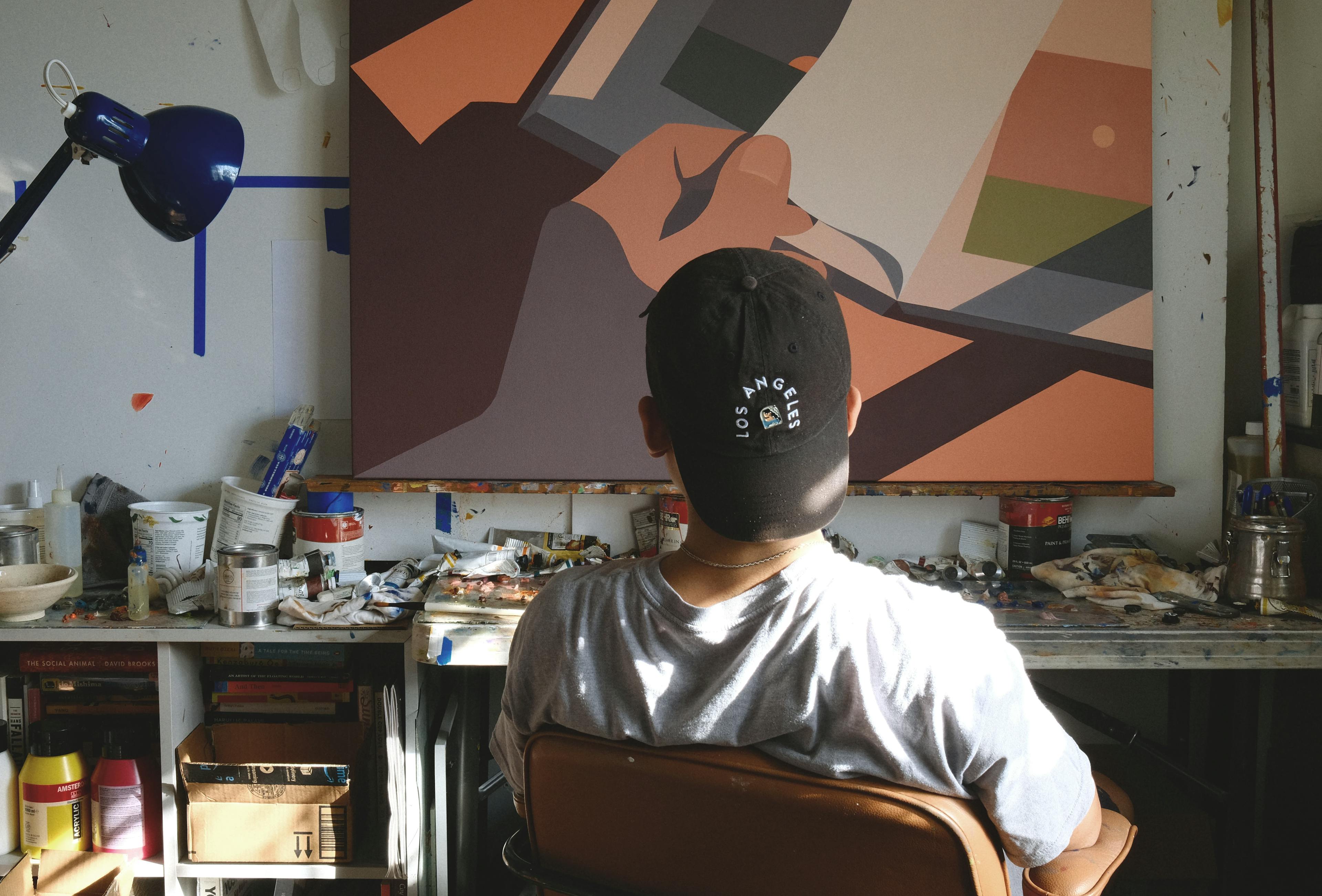 Artist Adrian Kay Wong, viewed from behind, sitting in front of a painting in his studio. 