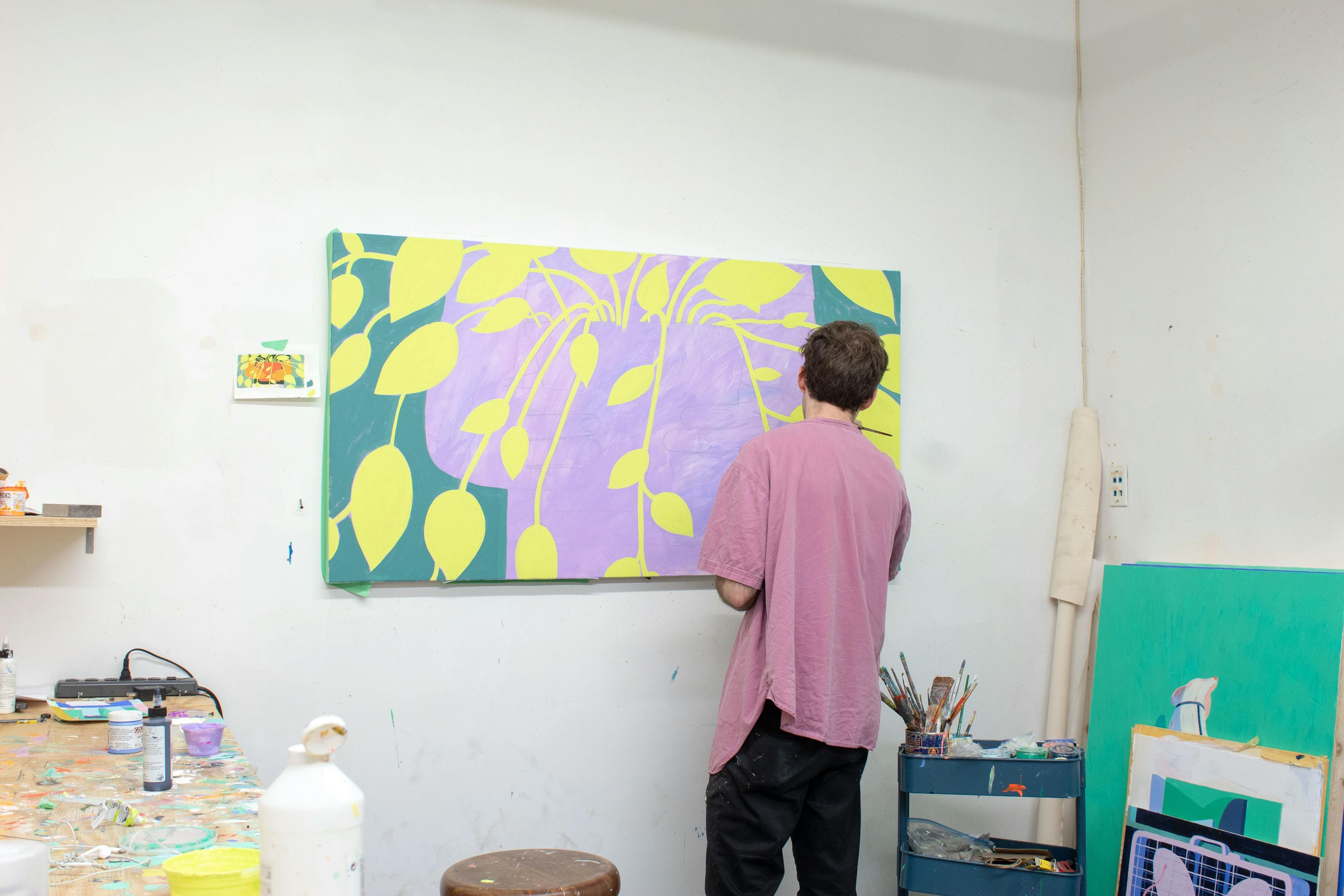 Artist Jackson Joyce painting a large botanical painting installed on a white wall in his studio.