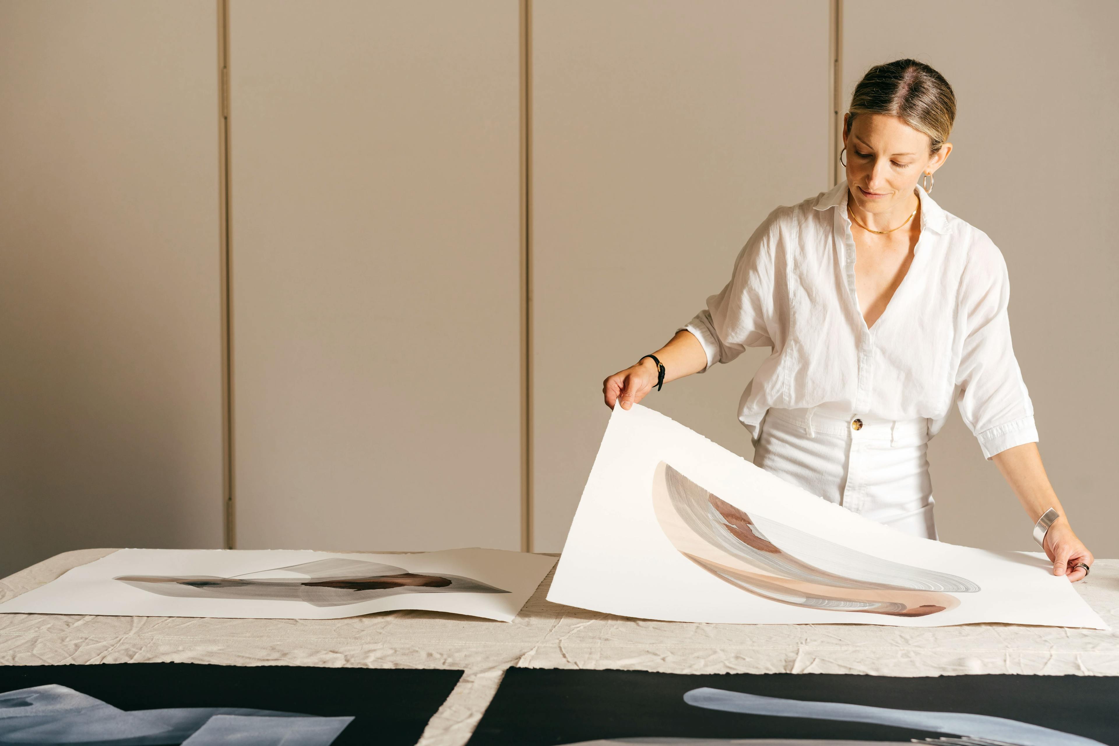 Artist Laura Naples dressed in white, standing at a table at MacArthur holding the corners of a gestural painting on white paper.