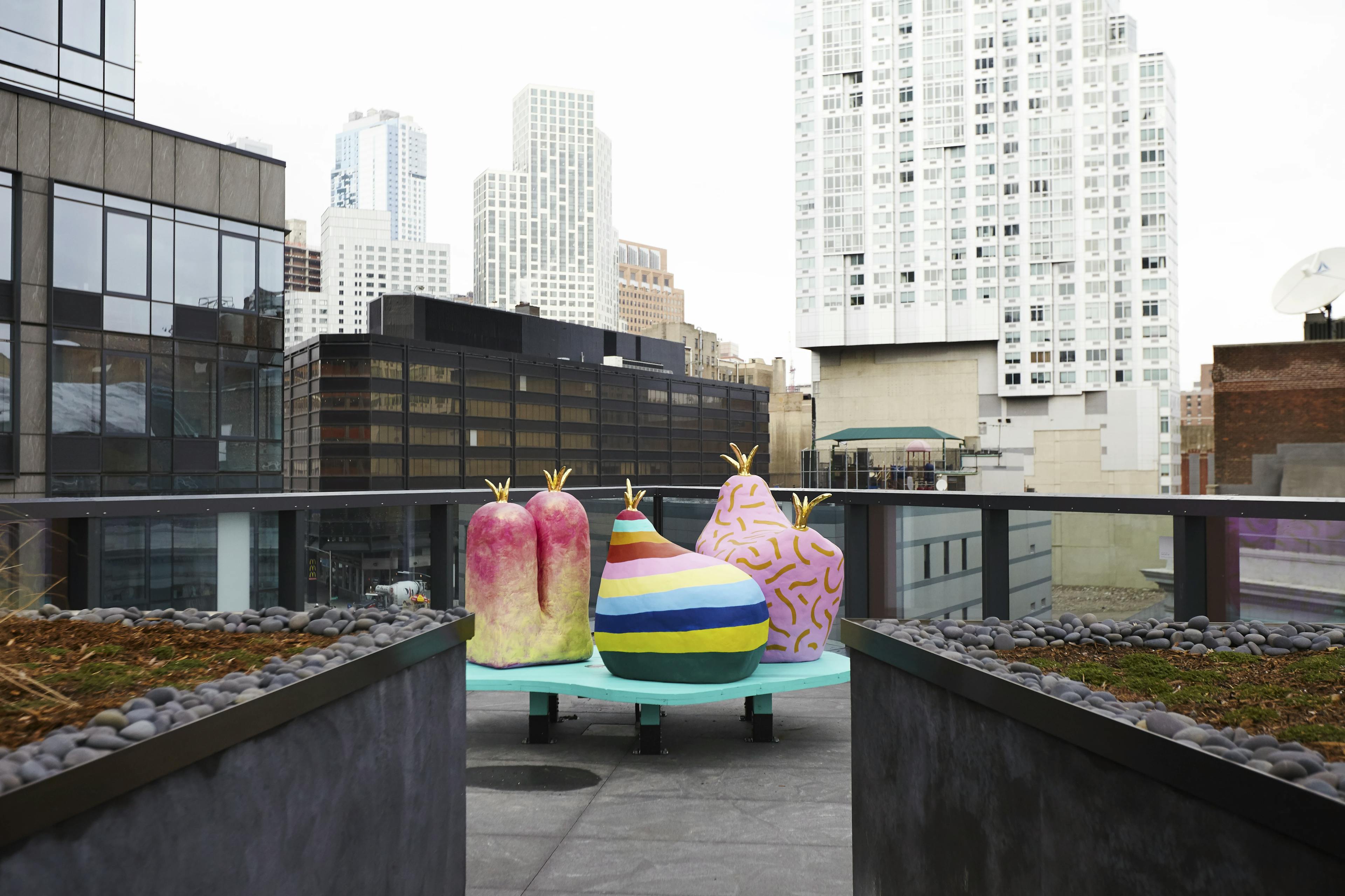 Colorful, large-scale sculptures by artist duo CHIAOZZA affixed to a blue platform on a building balcony in the middle of a cityscape with skyscrapers.