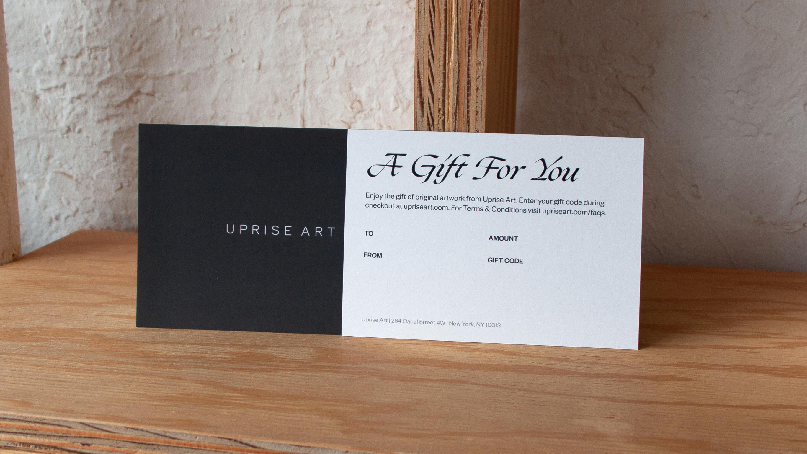 A gray Uprise Art gift card with text that says "A Gift For You" displayed on a wooden bookshelf at the Uprise Art gallery.
