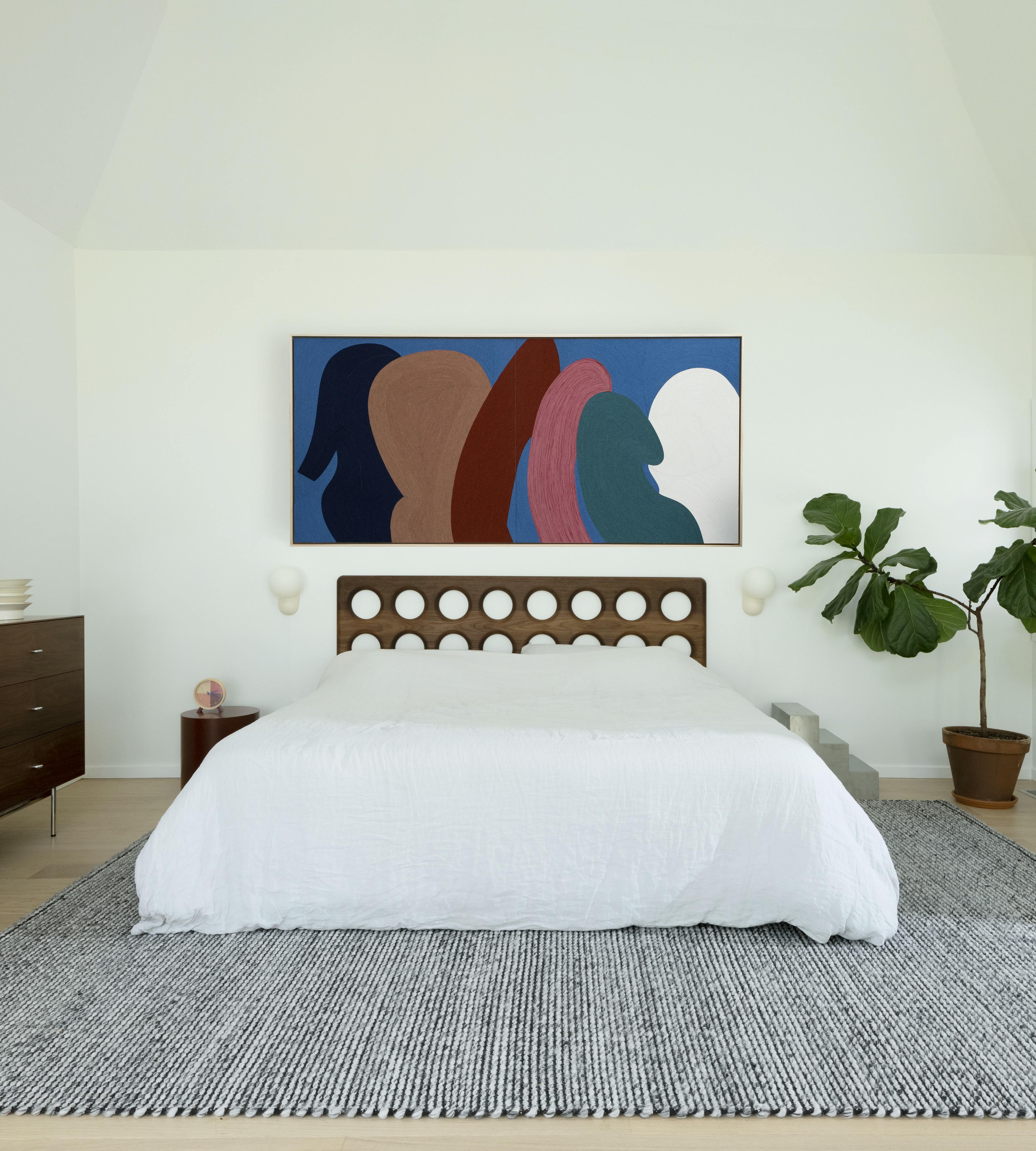 Large, abstract textile work made from yarn by artist Paola Rodriguez installed above a bed with white sheets and headboard with carved out circles.