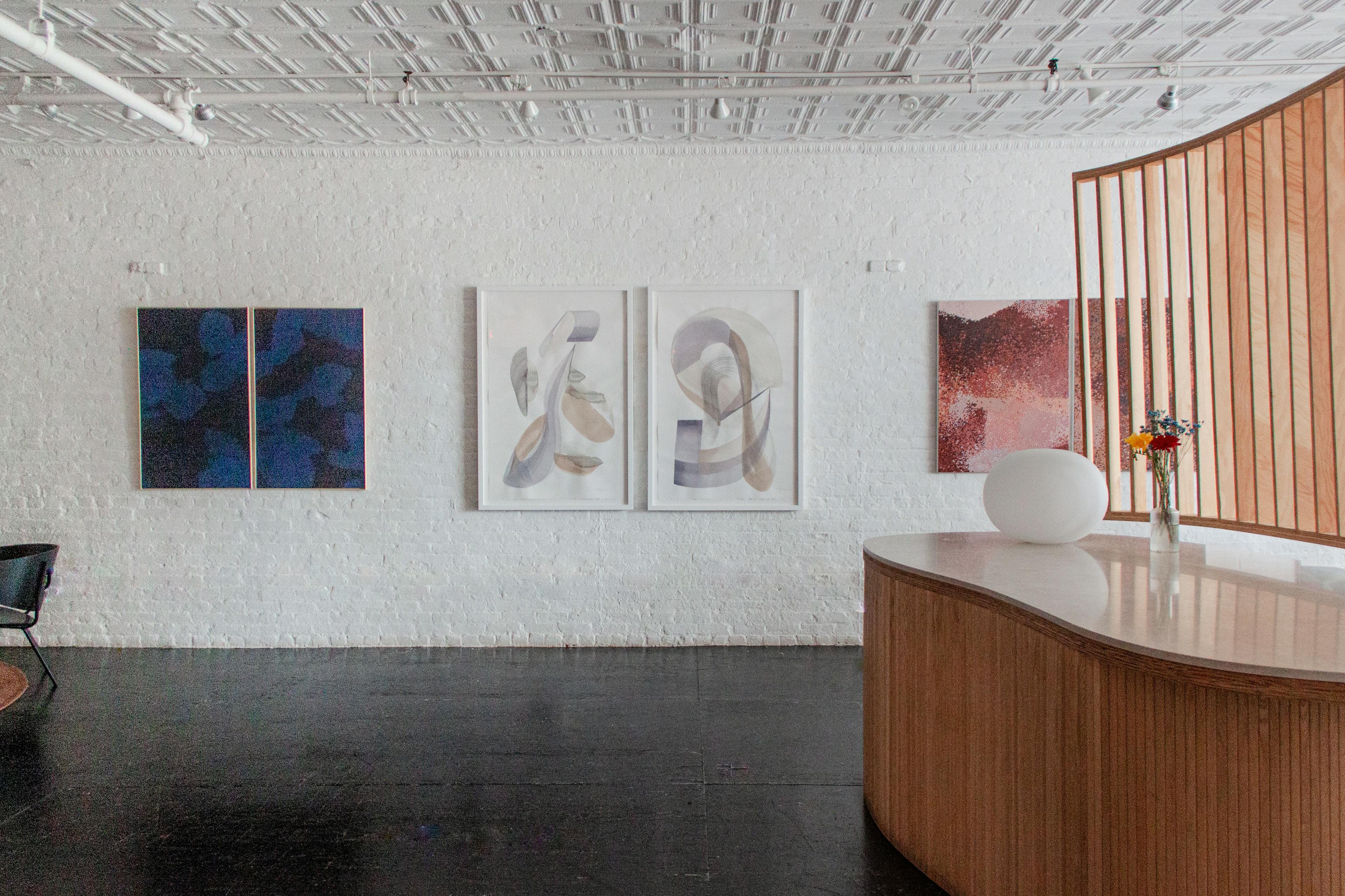 A dark blue diptych by artist Carrie Crawford, two abstract gestural paintings by artist Laura Naples, and a painting with repeating red and pink polka dots by Misato Suzuki, on a white brick wall at the Uprise Art showroom for the '23 collection.