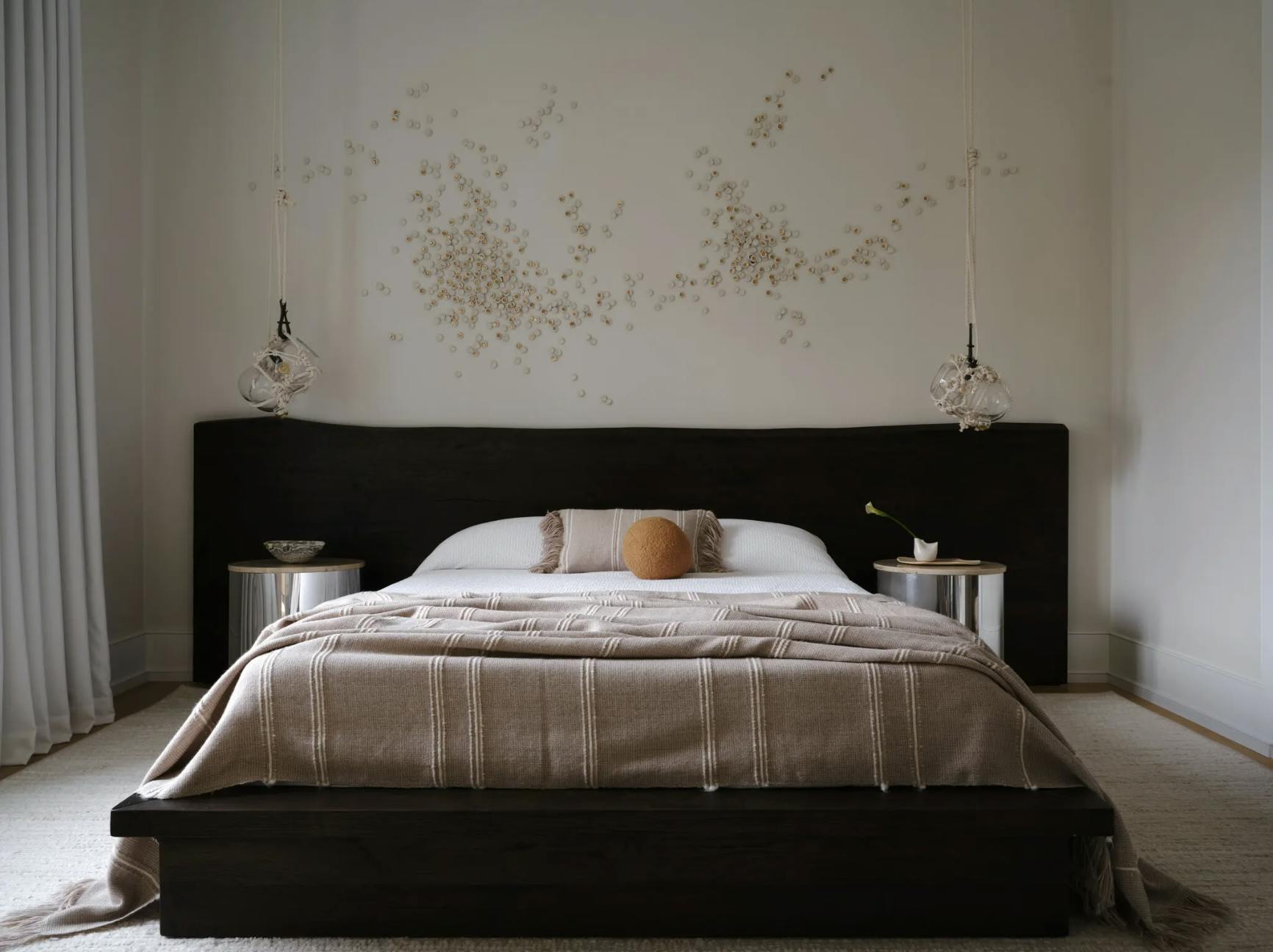 A custom installation by artist Christina Watka made from small pieces of mica positioned on a white wall above a bed with neutral sheets and a round orange pillow.
