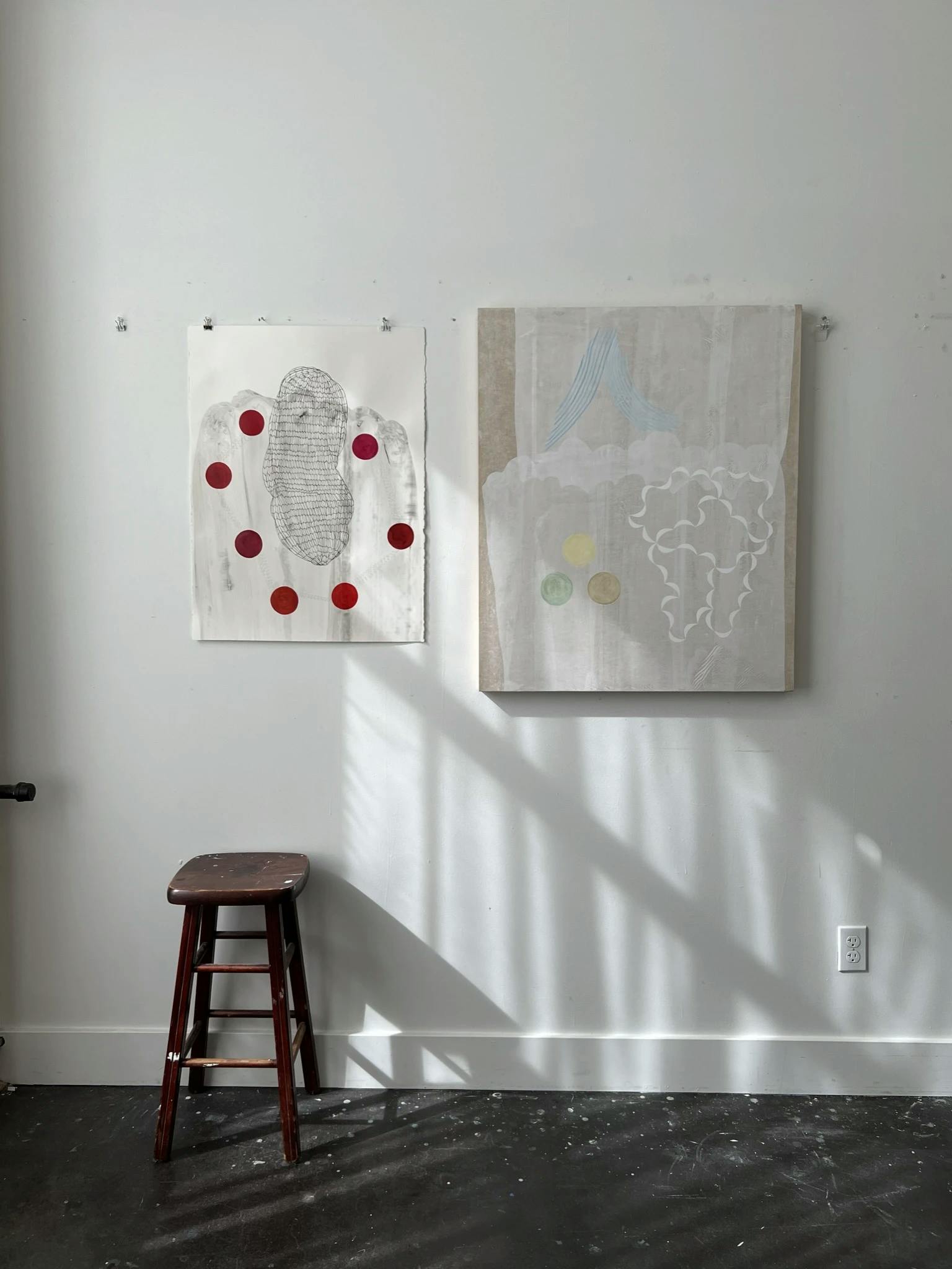 Two abstract paintings by artist Lydia Bassis installed on a white wall in her studio with a wooden stool on the left.