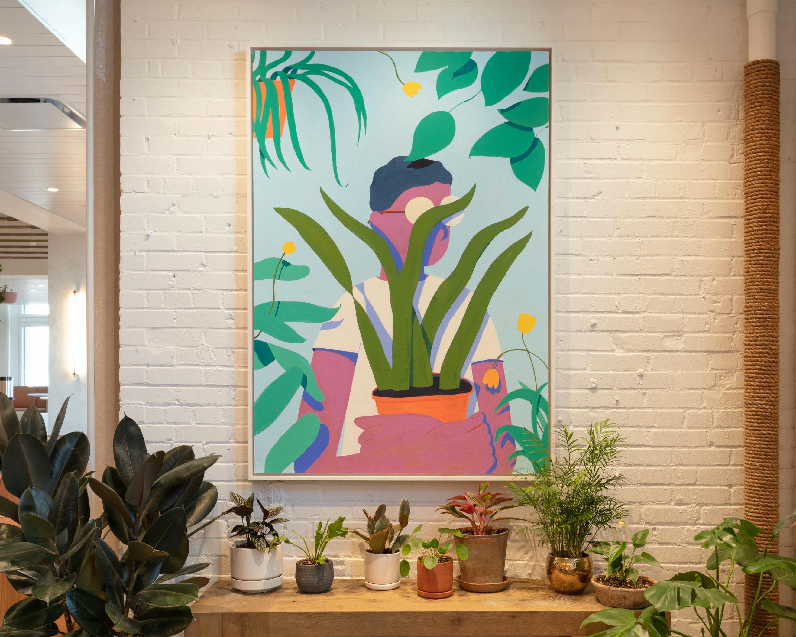 A painting by artist Jackson Joyce, depicting a person wearing glasses and standing among plants, installed above a table full of houseplants.